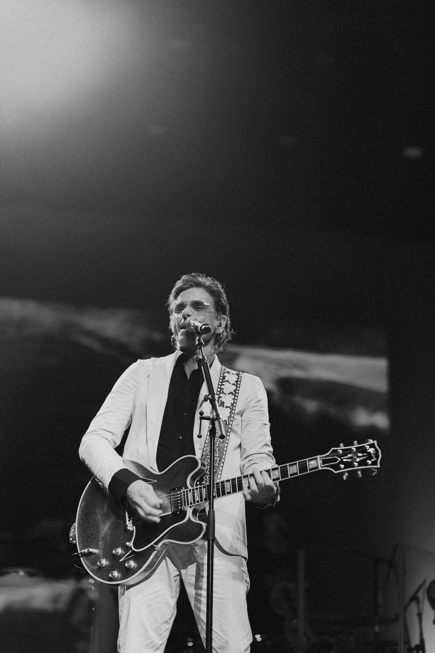 Musician in white jacket playing guitar on darkened stage during performance.