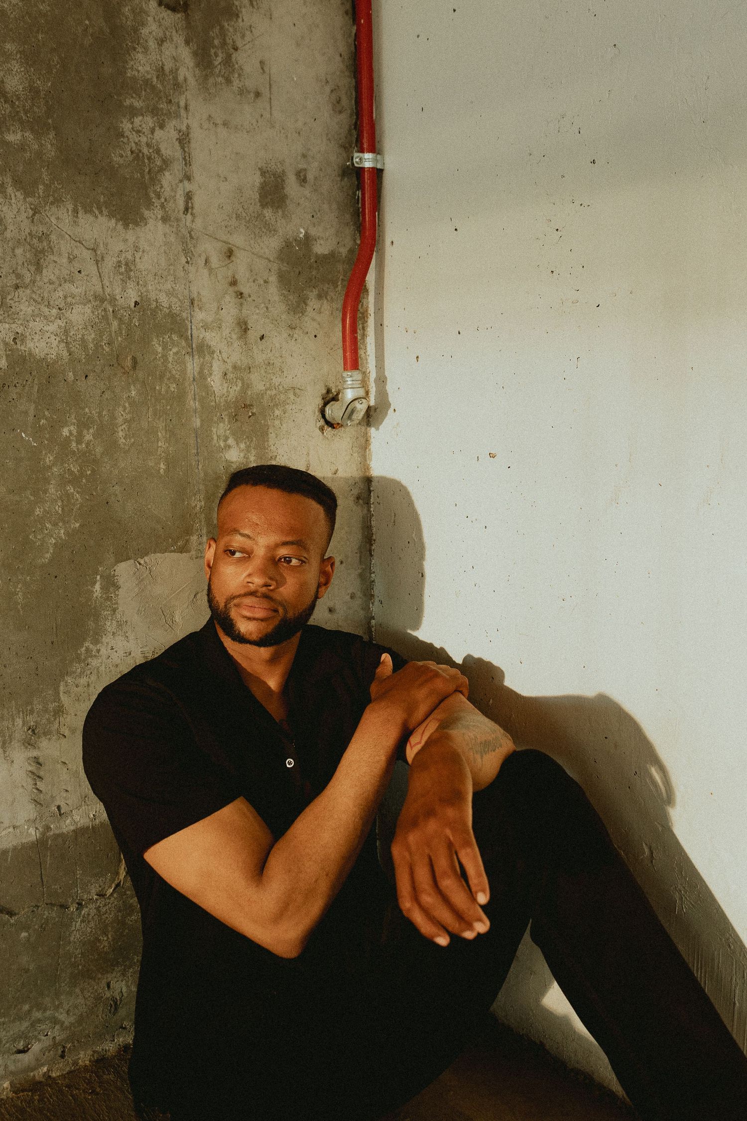 A man in a black shirt sits against a concrete wall with a red pipe, posing confidently in moody lighting.