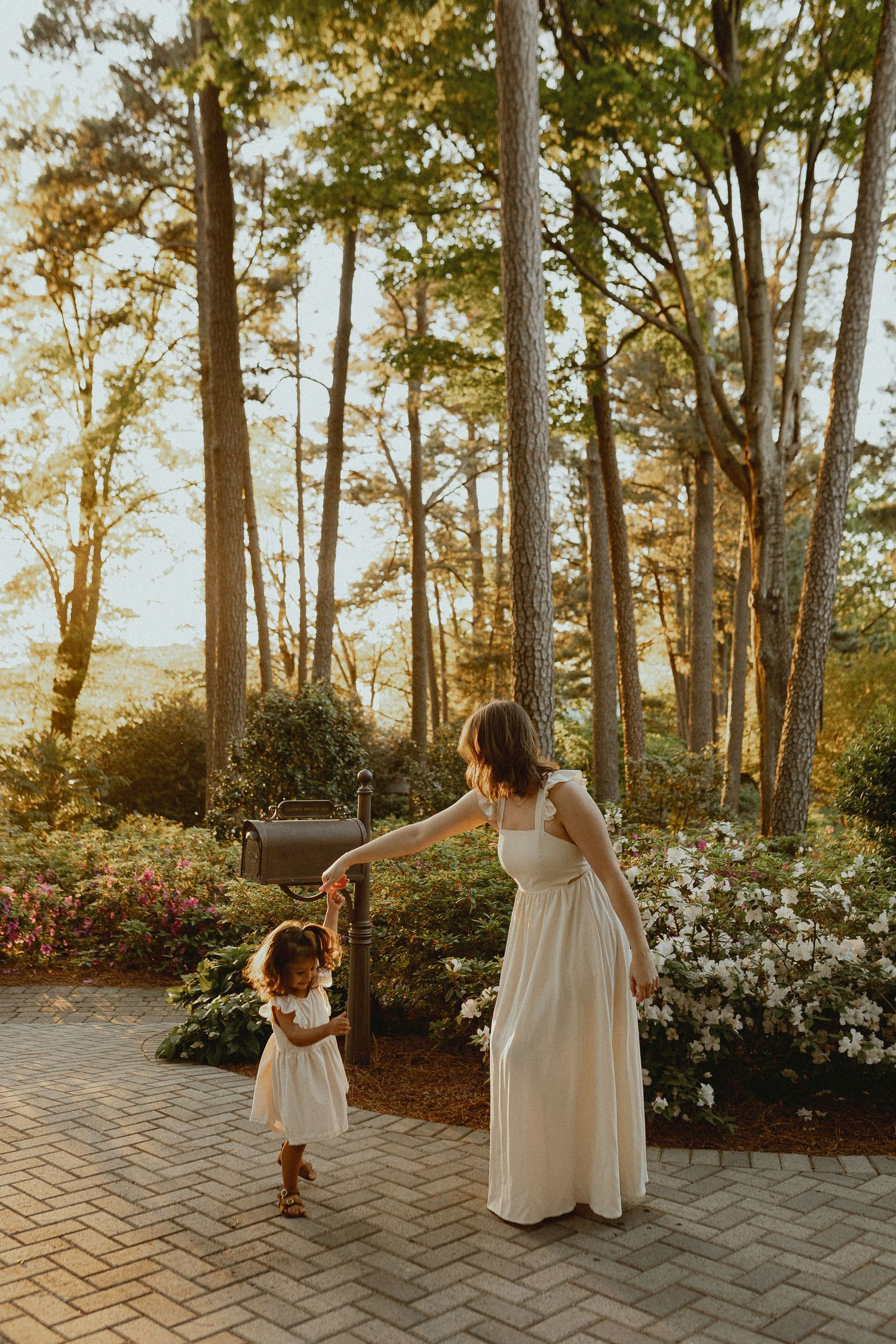 A white-dressed figure twirls playfully on a stone patio surrounded by tall trees and flowering bushes at sunset.