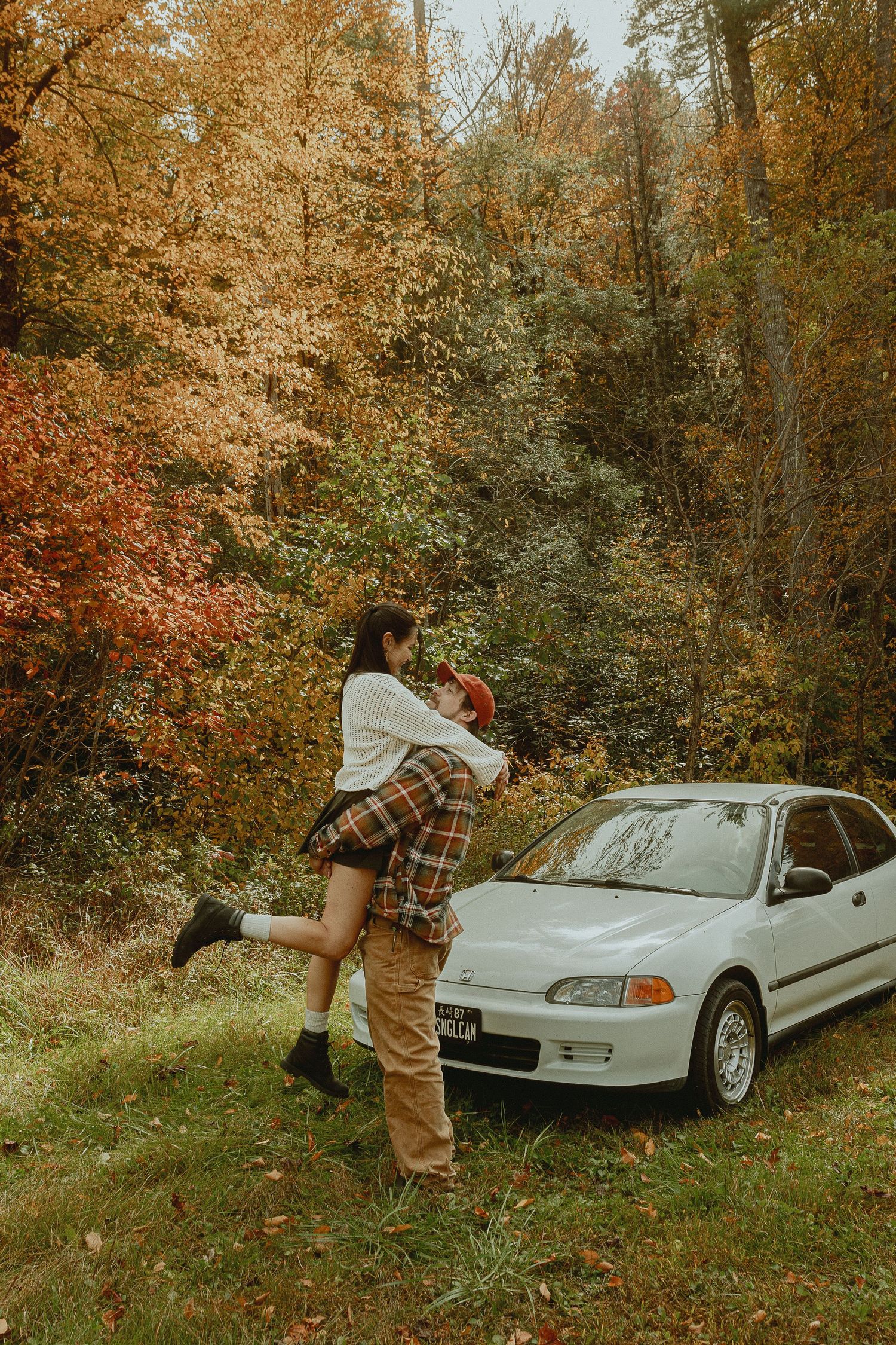 Couple embracing by a white Honda Civic amid vibrant autumn foliage in a forest setting.