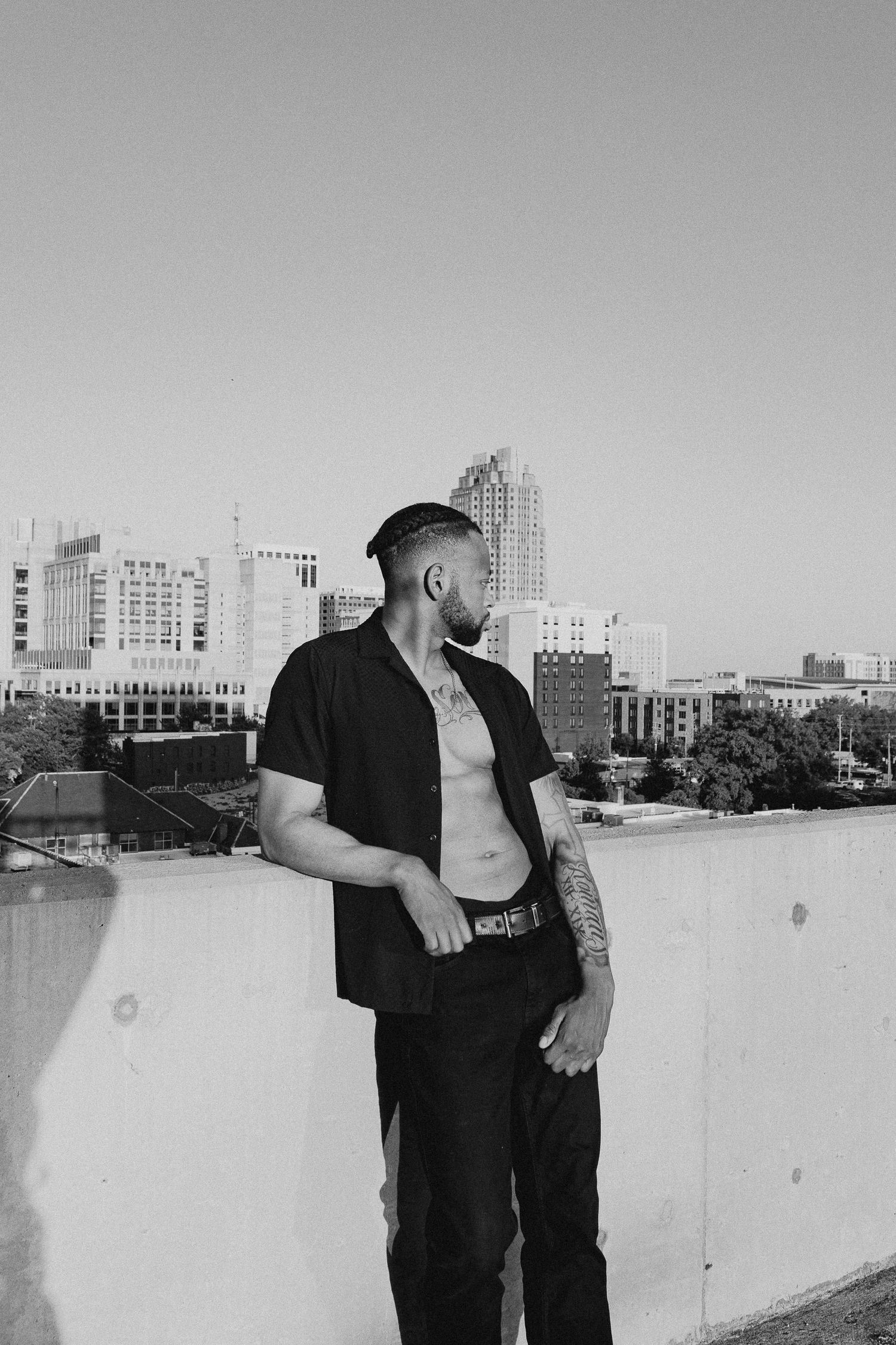 Black and white photo of tattooed shirtless man in open shirt posing on rooftop with city skyline background.