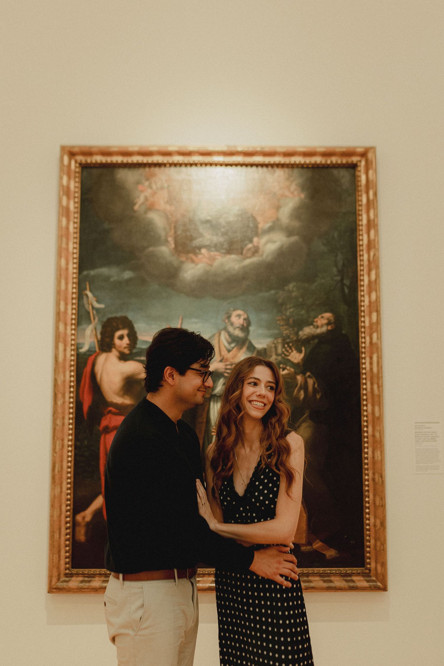 Couple smiling together in front of a large classical religious painting in an art museum gallery.