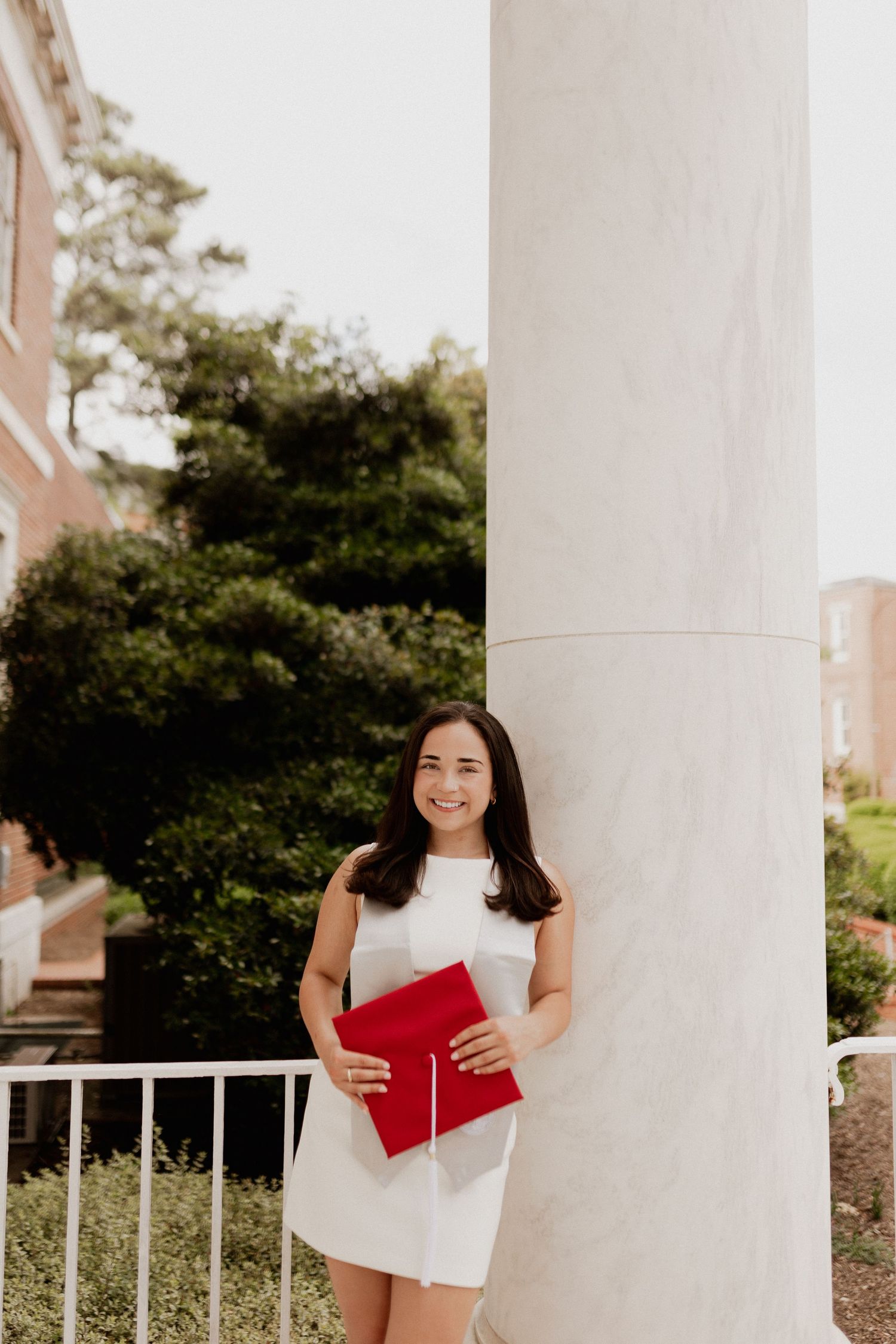 A person poses with a red folder in front of white columns on a sunny day.