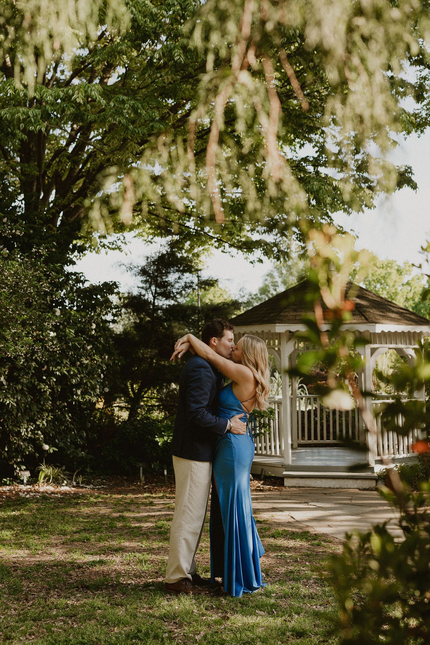 Couple sharing a romantic kiss in a lush garden near a white gazebo, woman in blue gown, man in navy suit.