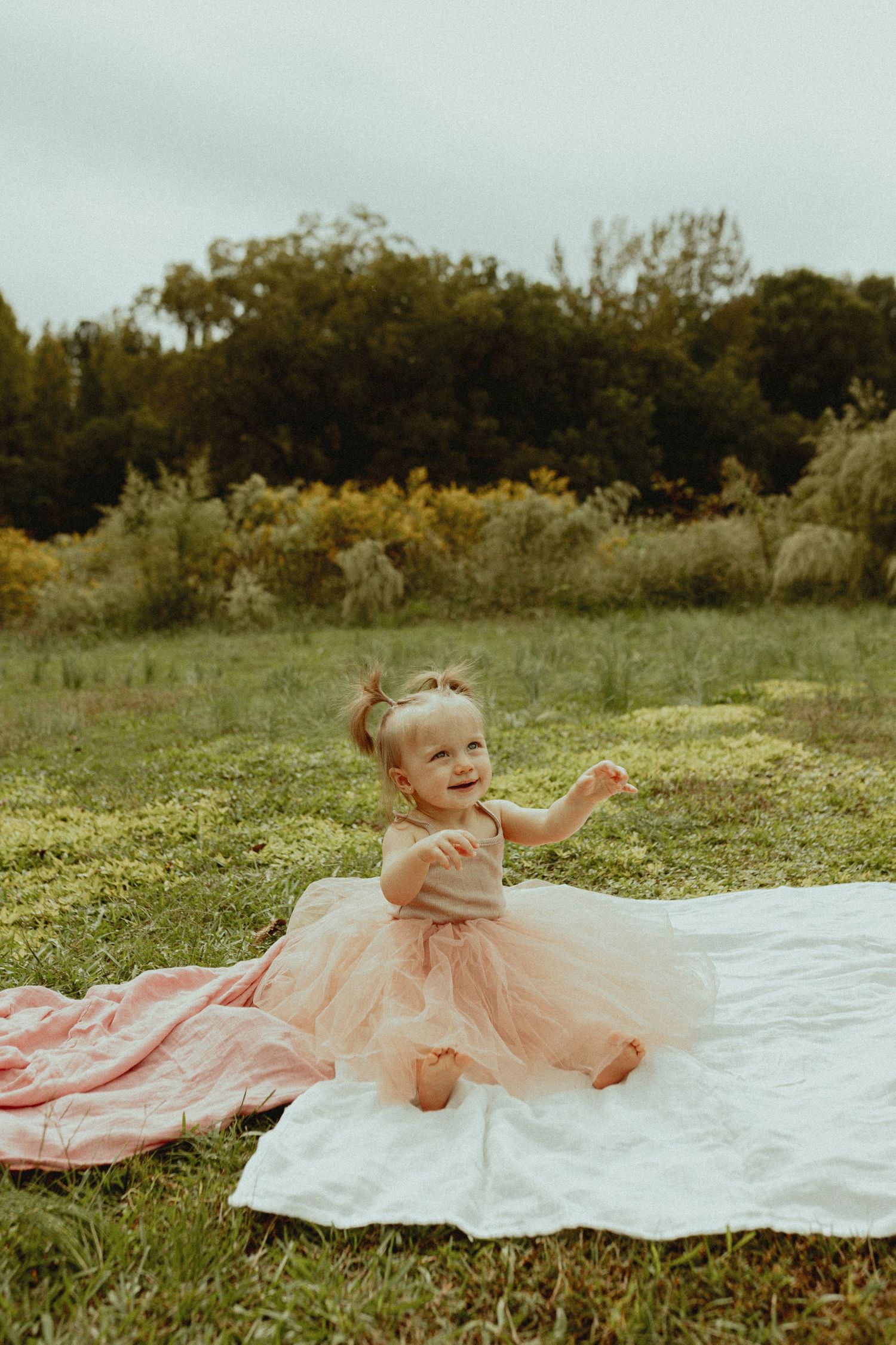 A baby wearing a pink tulle dress smiles while sitting on a white blanket.