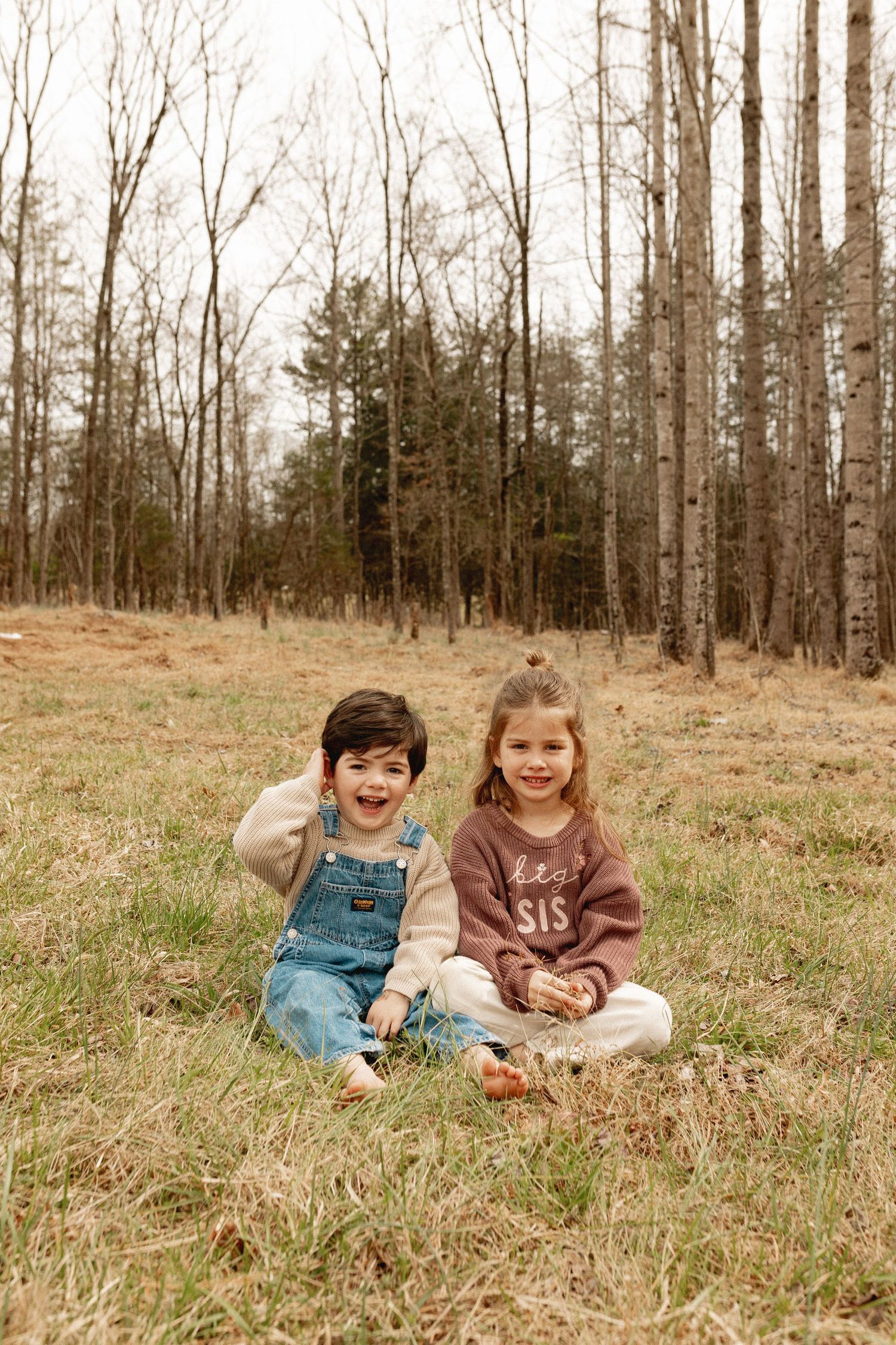 Two young children sitting on grass in a woodland clearing, one in denim overalls, the other in a brown SIS sweatshirt.