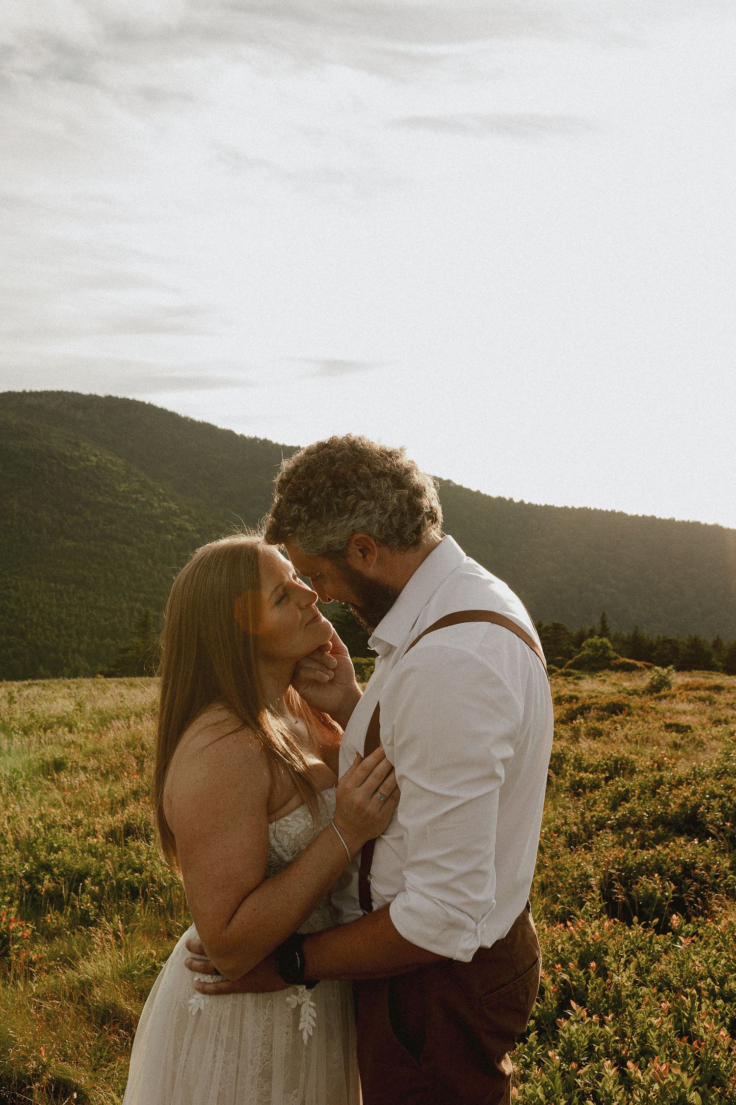 Couple sharing a romantic moment at golden hour in a mountain meadow, bride in white dress, groom in suspenders.
