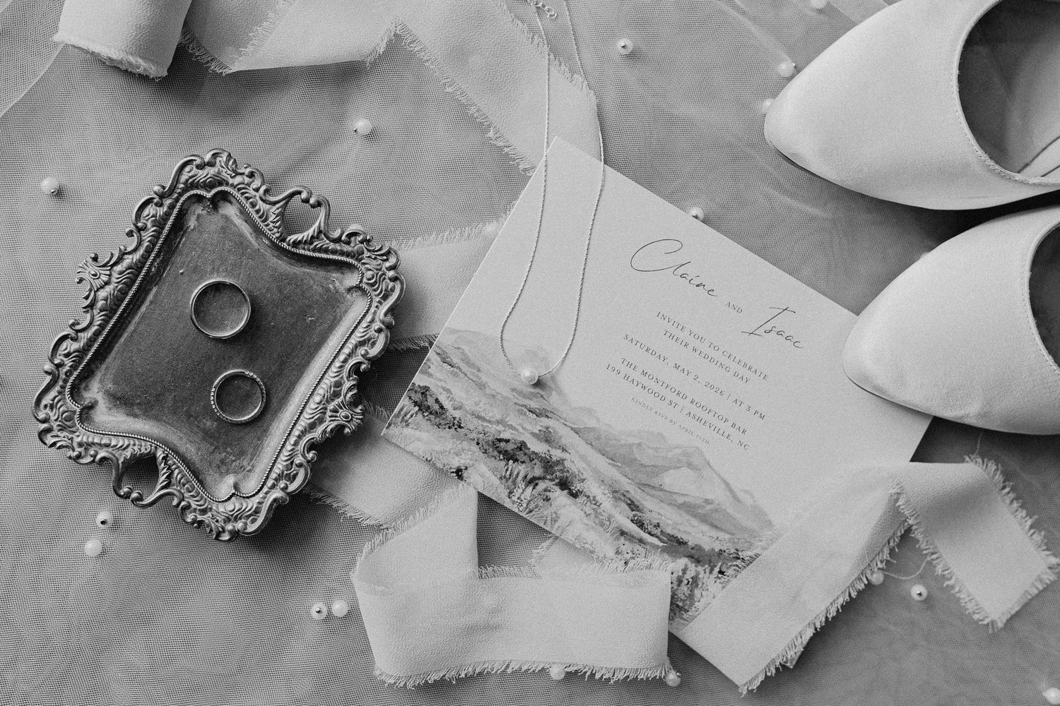 Black and white wedding flat lay with rings, invitation, bridal heels, pearls, and ribbon on soft fabric.