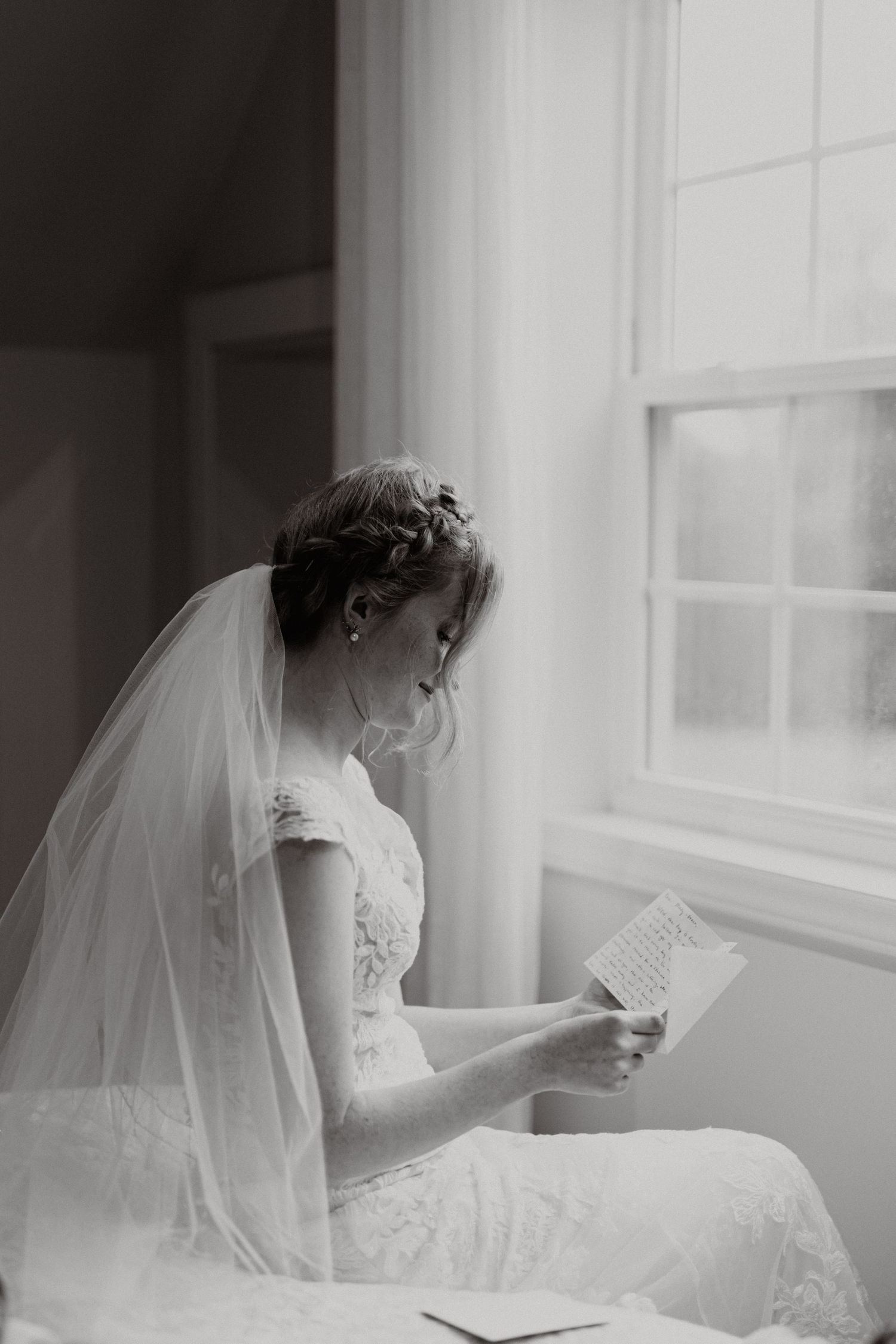 Black and white photo of bride in veil gazing thoughtfully out window.