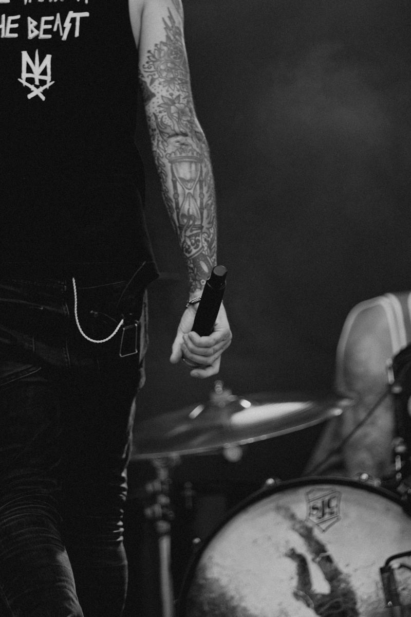 Close-up of tattooed arm holding drumstick against dark background with band logo visible.