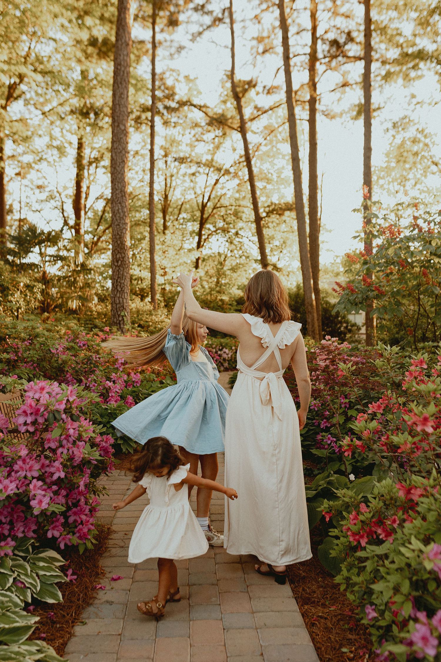 Three figures in flowing dresses dance along a garden path lined with vibrant pink azalea bushes and tall trees.