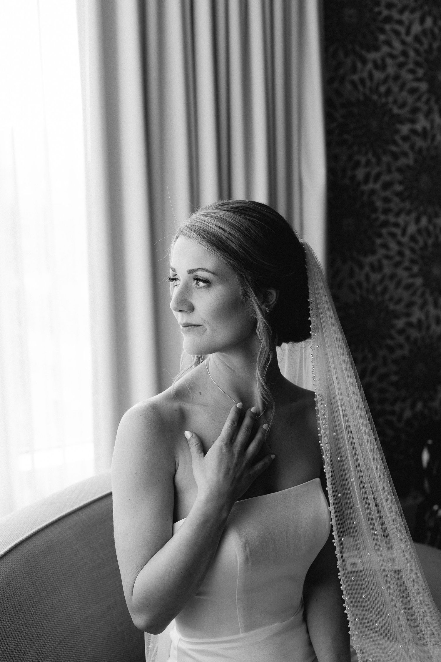 Black and white portrait of a bride in a strapless gown and pearl veil gazing out a window on her wedding day.