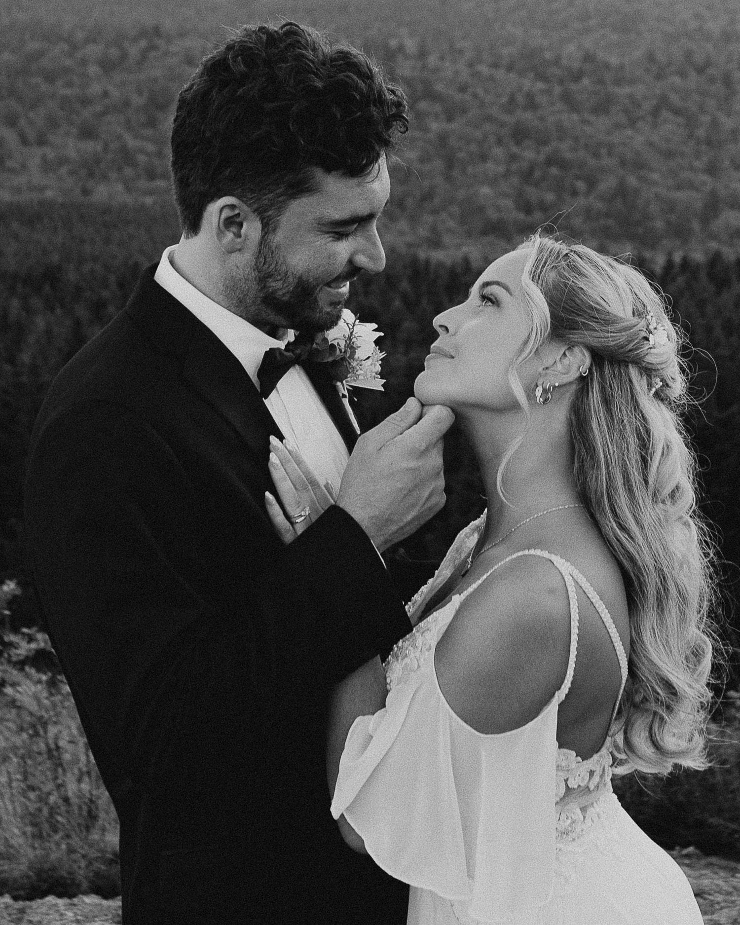 Black and white wedding photo of bride and groom gazing at each other outdoors with forest backdrop.