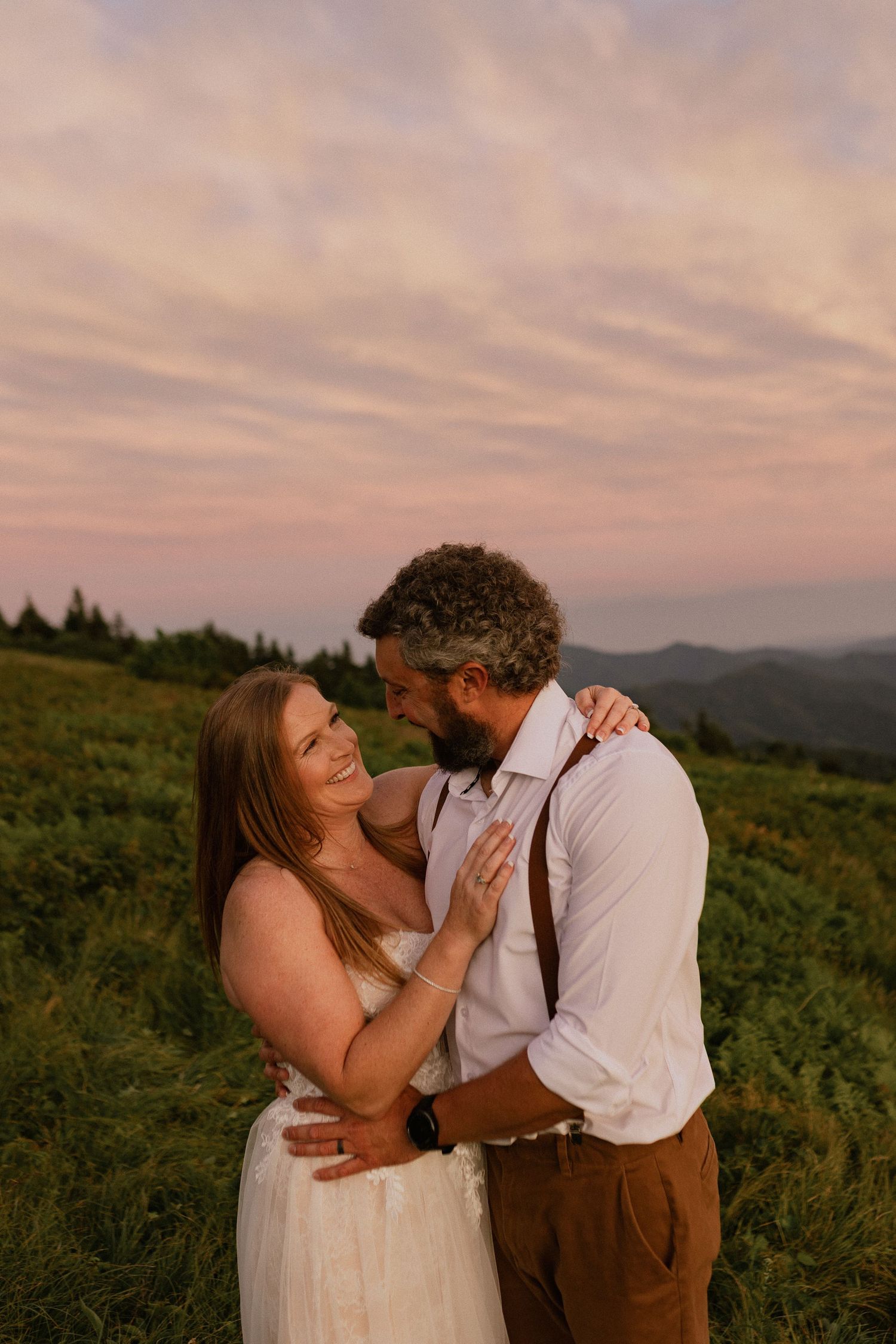 Couple embracing at sunset on a mountain meadow, woman in white dress, man in suspenders, pink sky backdrop.