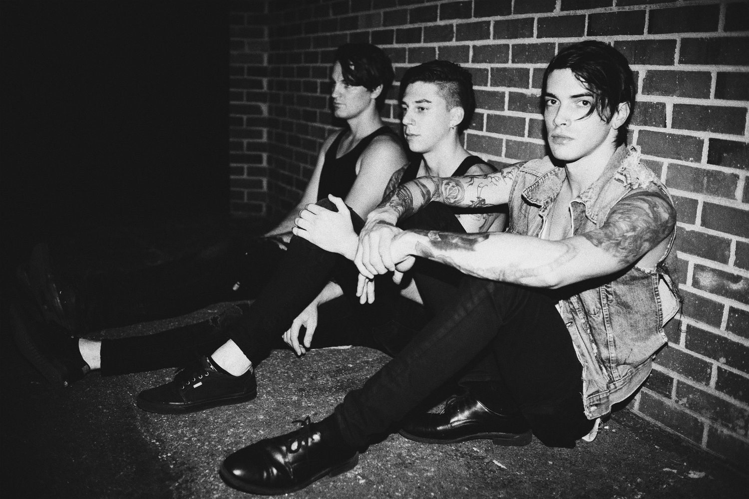Three people sitting casually against brick wall in black and white photography.