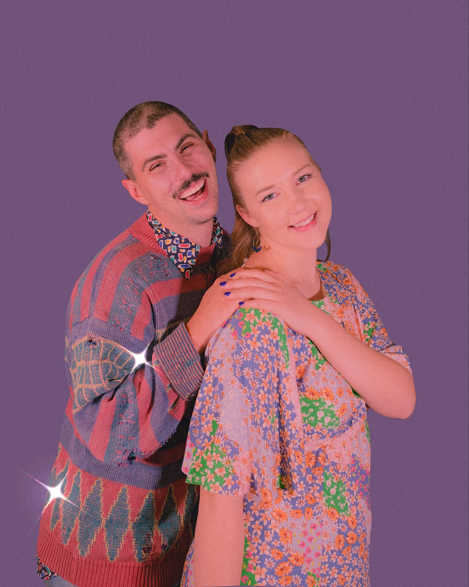 Couple poses playfully against purple background wearing colorful vintage sweater and floral top.