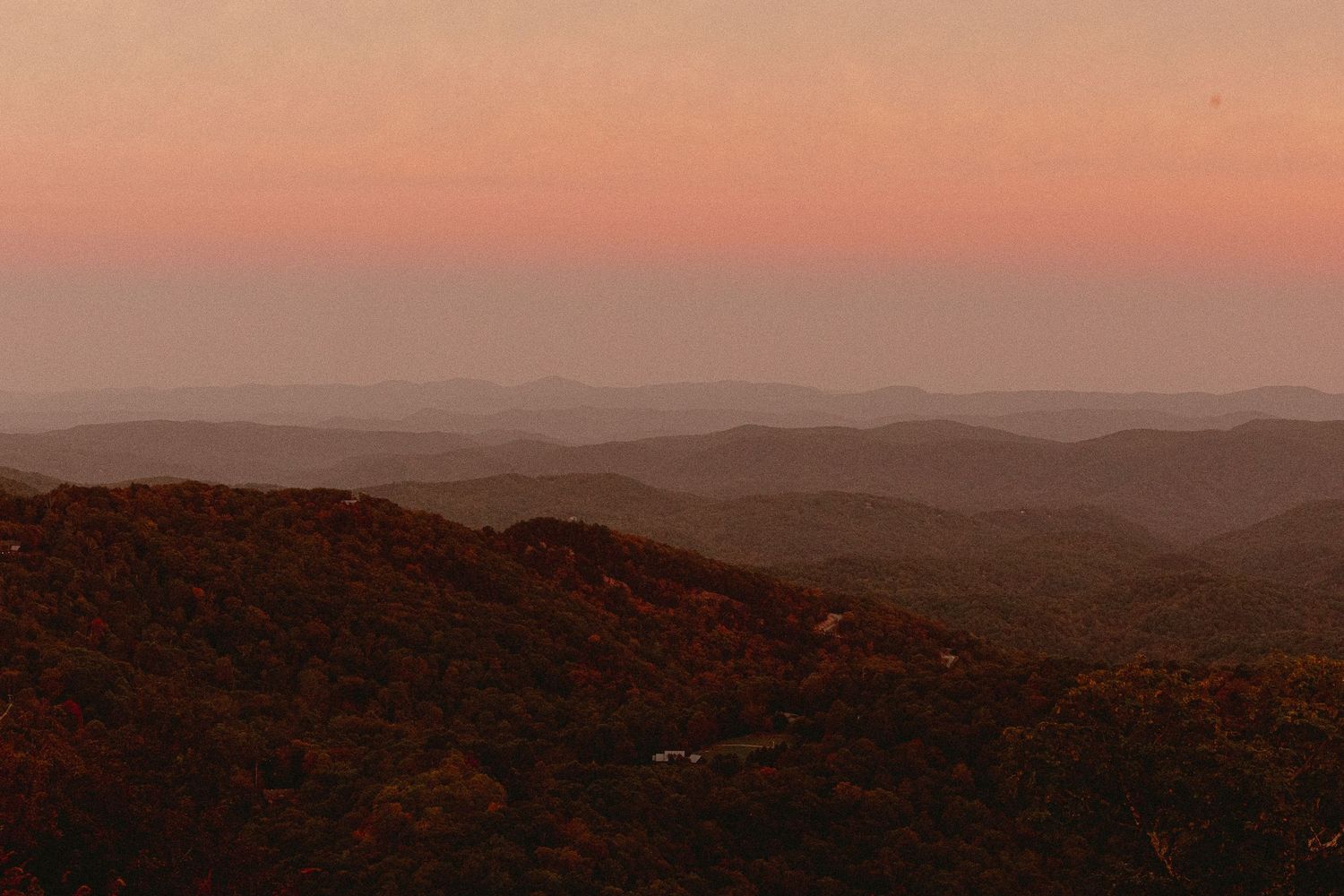 Aerial view of rolling mountains covered in autumn foliage at sunset with warm pink and orange sky.
