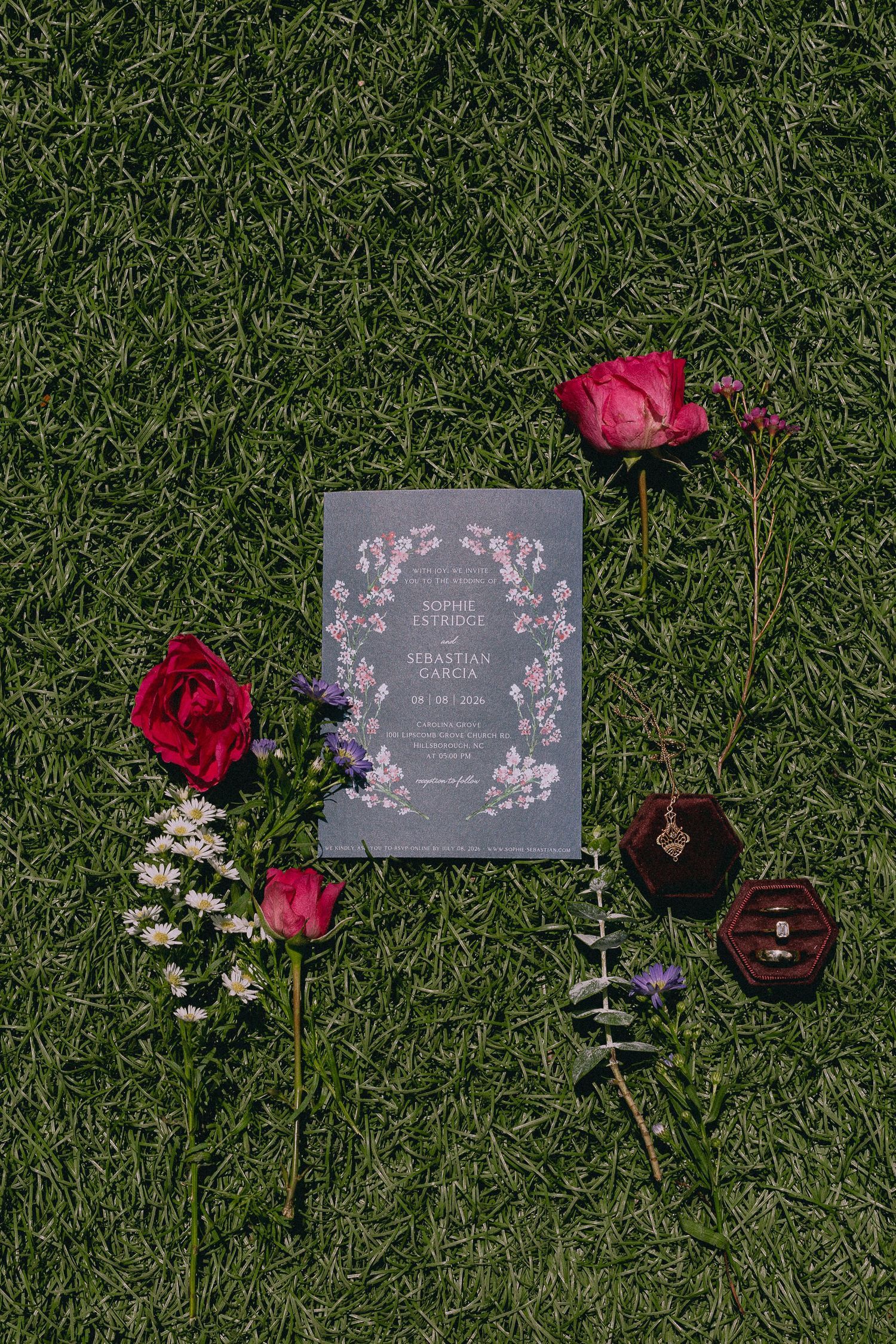Wedding invitation suite on green grass with red roses, purple flowers, eucalyptus, and a velvet ring box.
