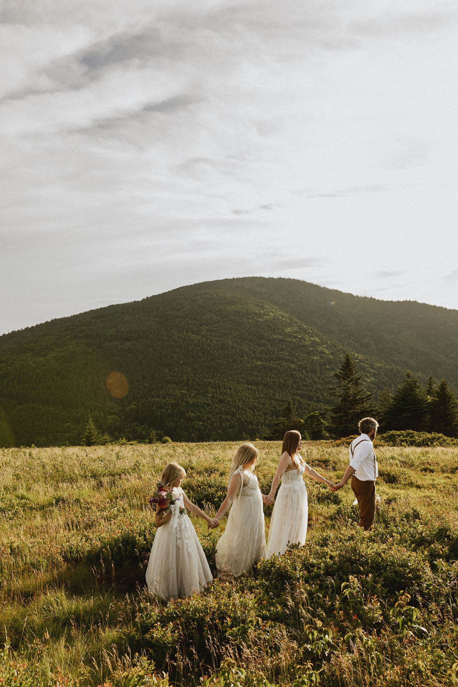 Children in white dresses and a boy in suspenders walk hand-in-hand through a mountain meadow at golden hour.