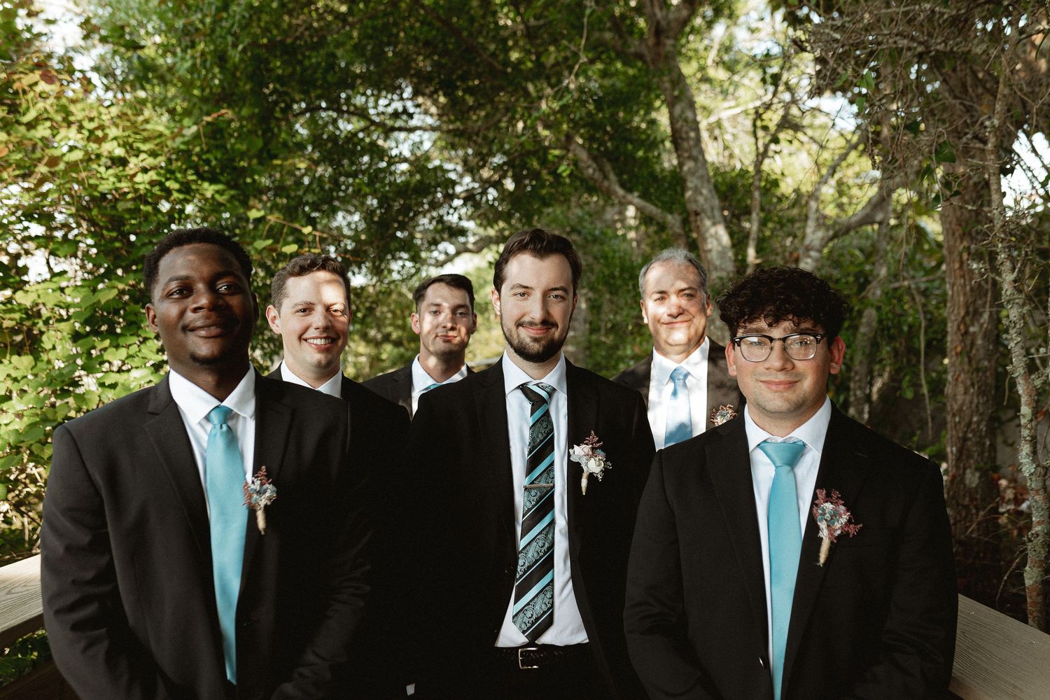 Groomsmen in dark suits with turquoise ties pose together outdoors for a wedding photo.