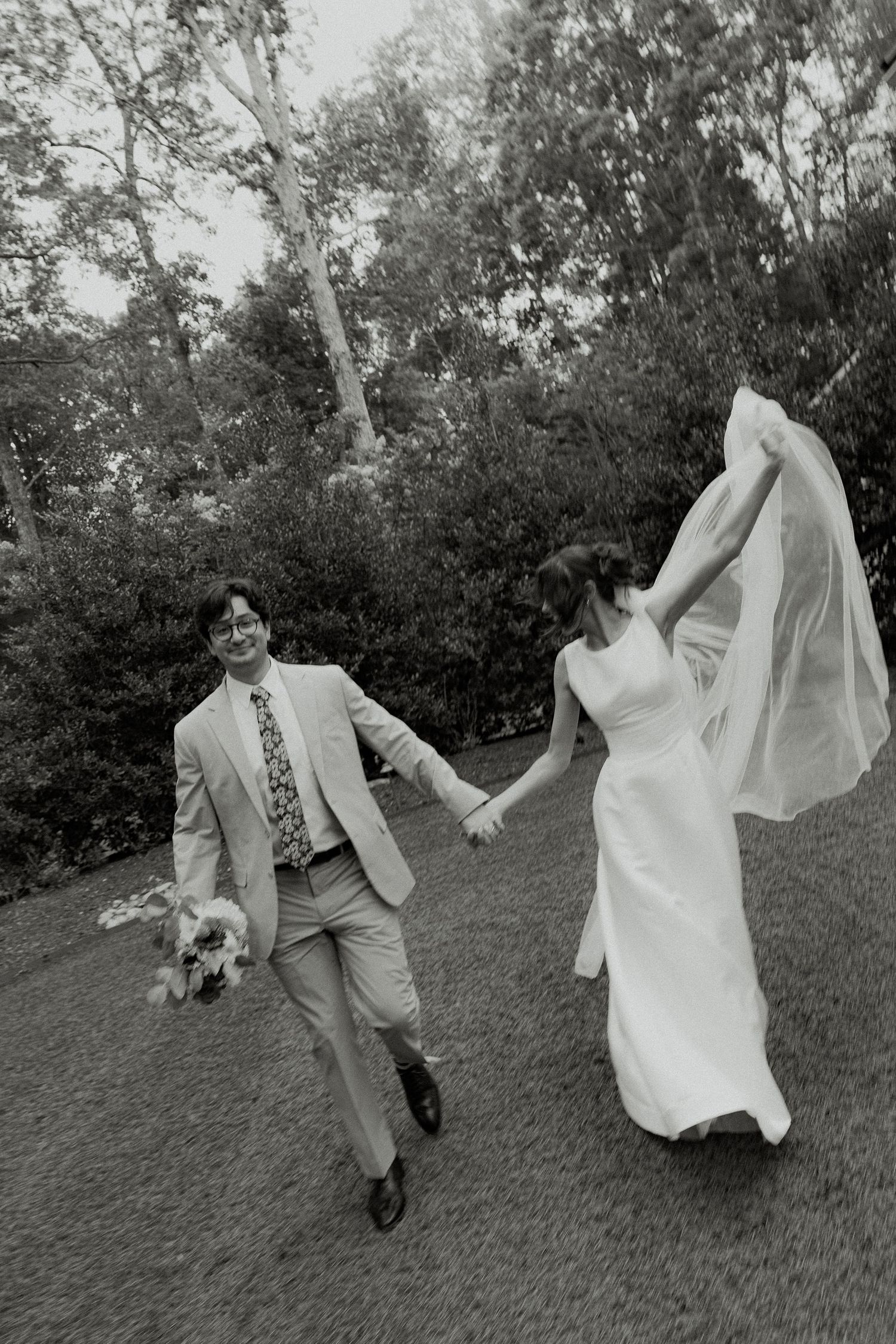 Black and white photo of a bride and groom holding hands, running through a garden, her veil flowing in the wind.