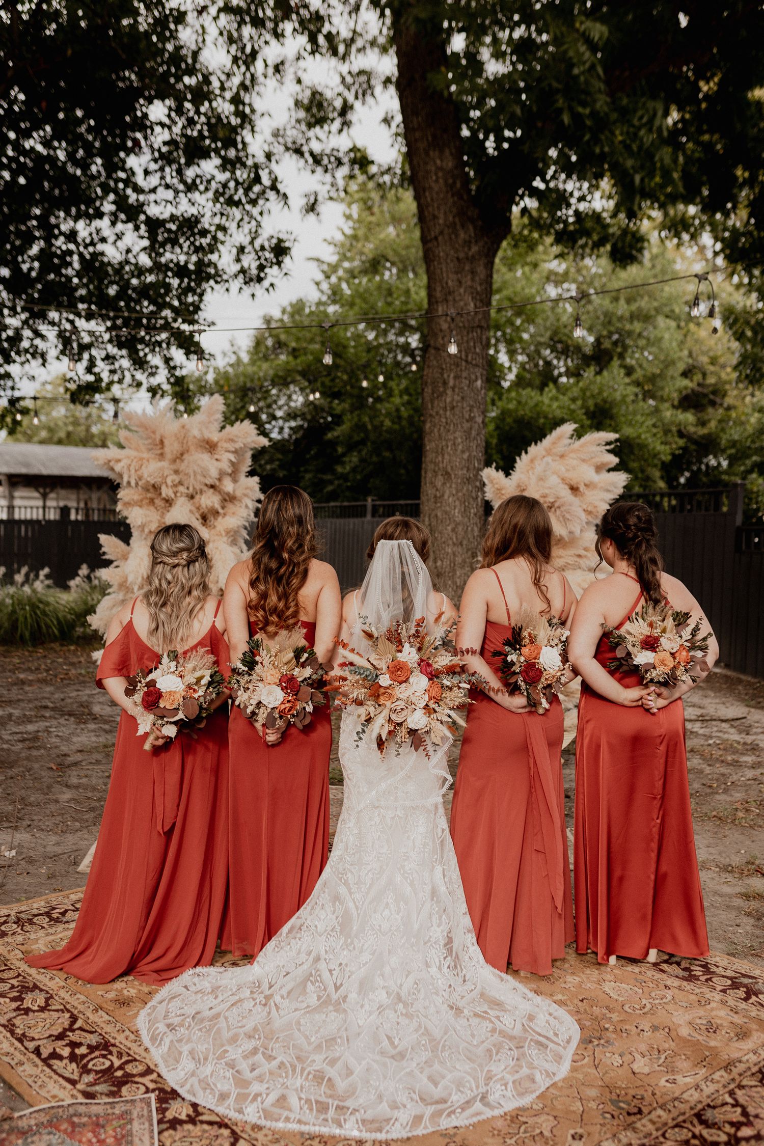 Bridesmaids in rust-colored dresses stand together with a bride in a white lace gown against a natural outdoor backdrop.