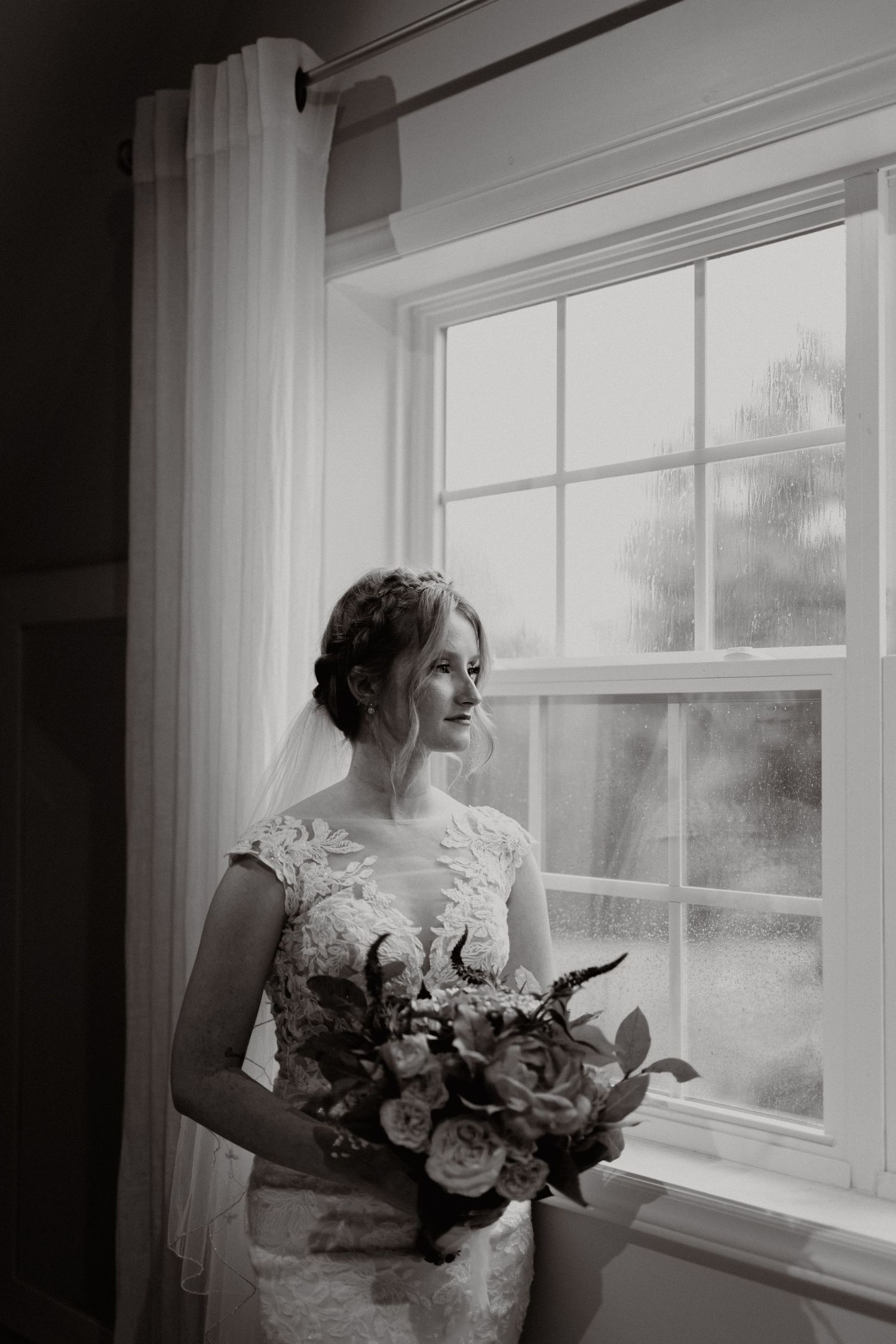 A person in a wedding dress gazes out a bright window while holding a floral bouquet.