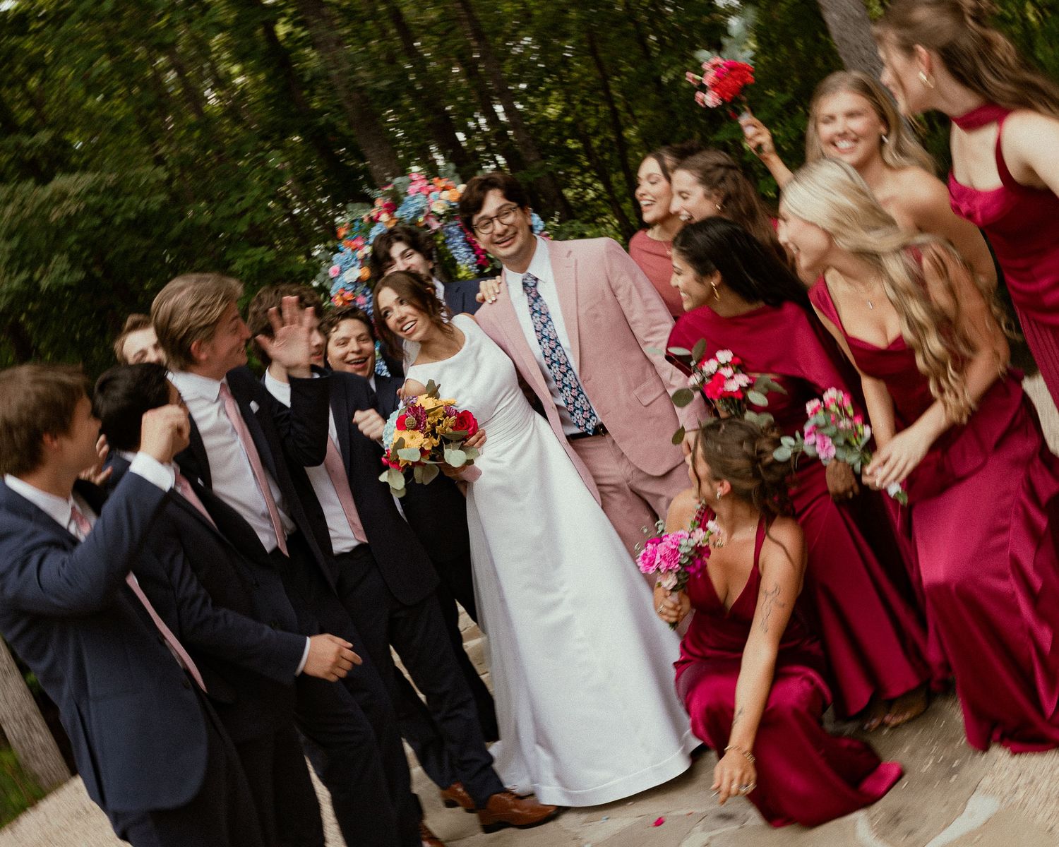 Bride and groom celebrate with wedding party, bridesmaids in red dresses and groomsmen in dark suits outdoors.