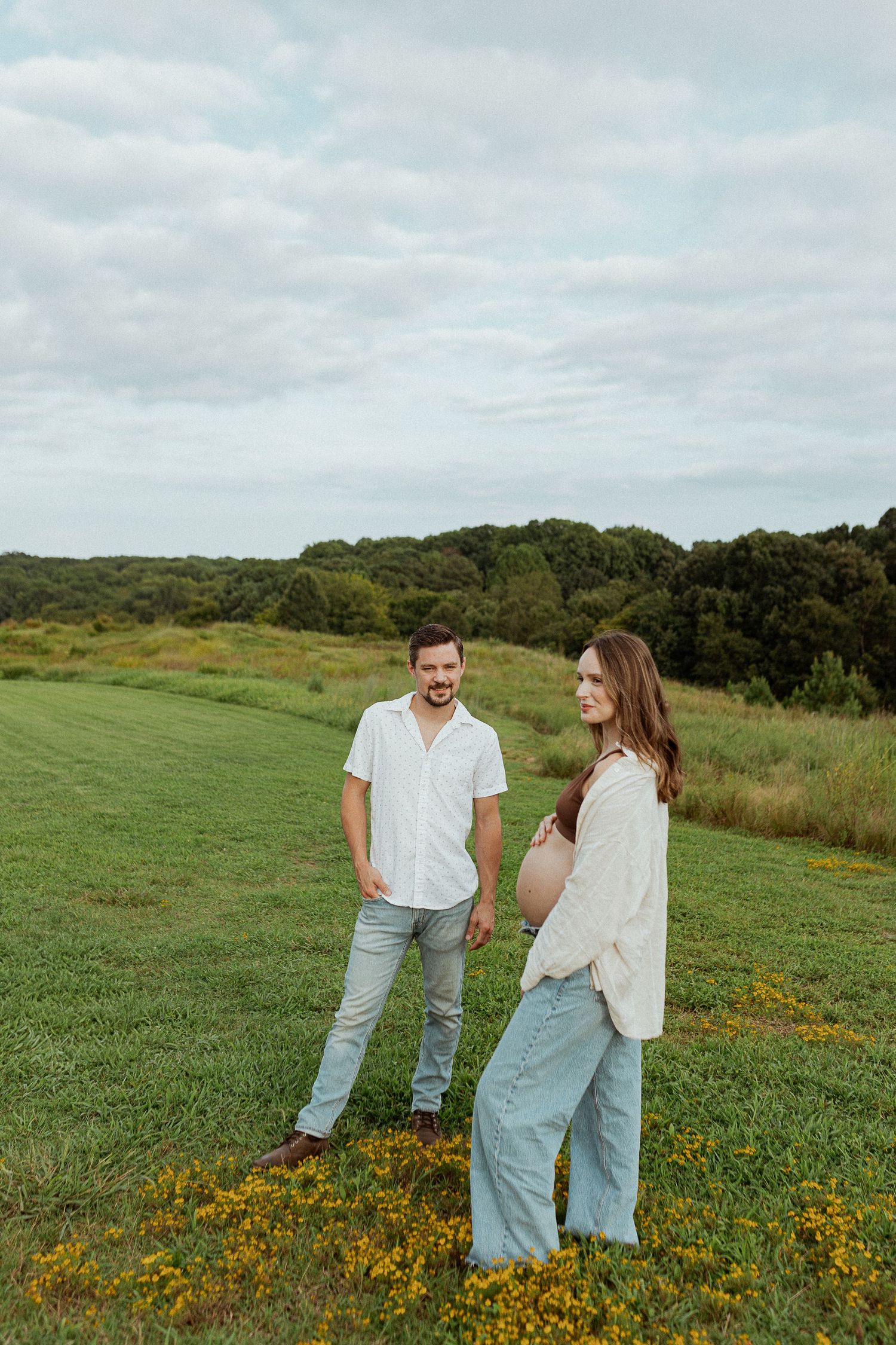 Couple standing in field of yellow wildflowers with green hills and cloudy sky in background.