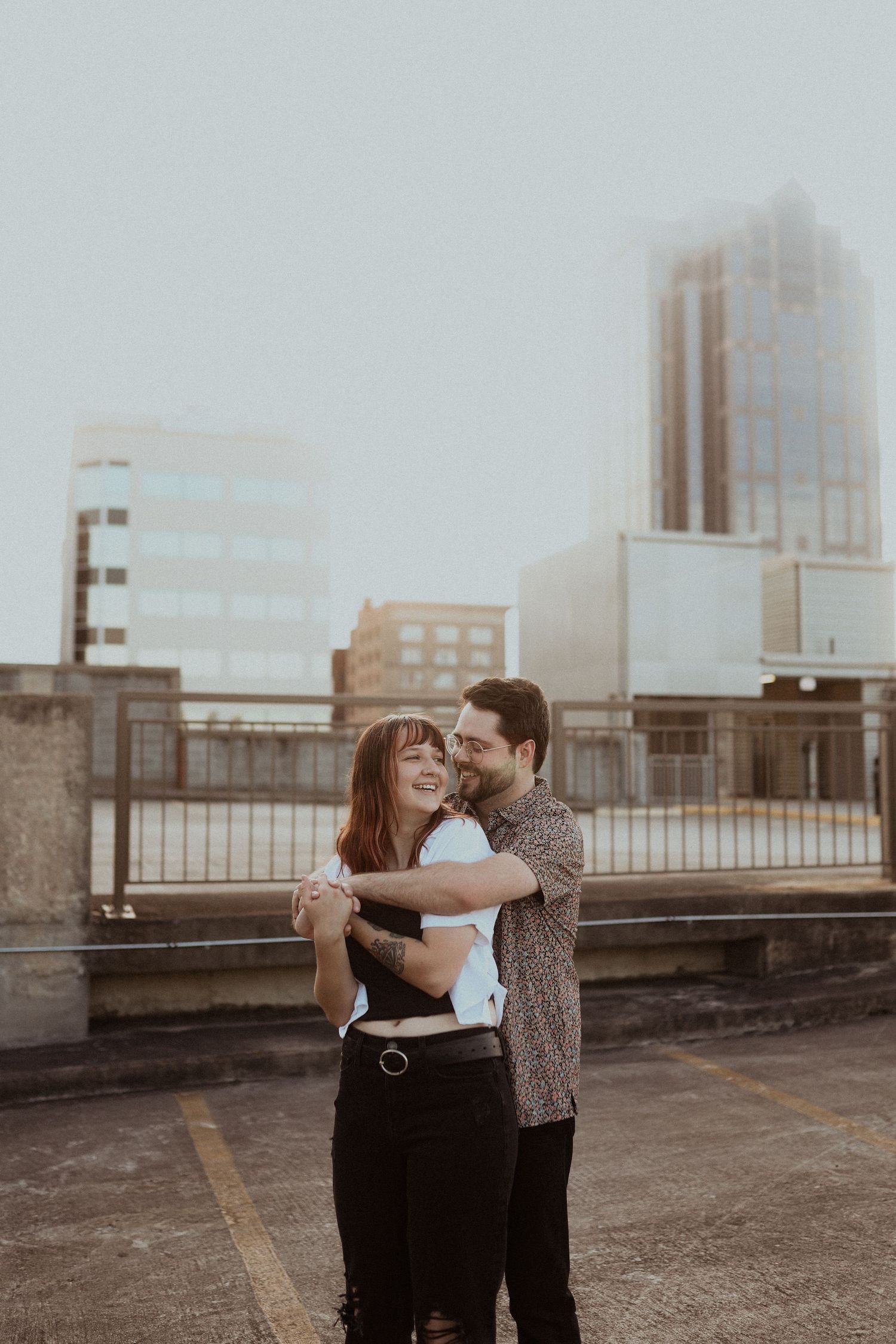 A couple shares a romantic moment while dancing on an urban rooftop with city buildings in the background.