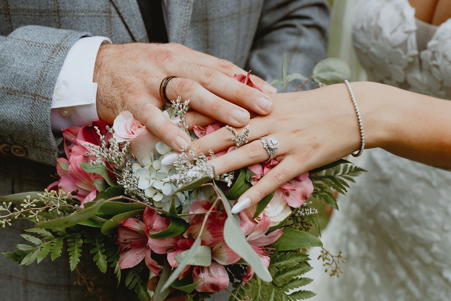 Bride and groom hands resting on pink floral wedding bouquet, showing wedding rings and elegant manicured nails.