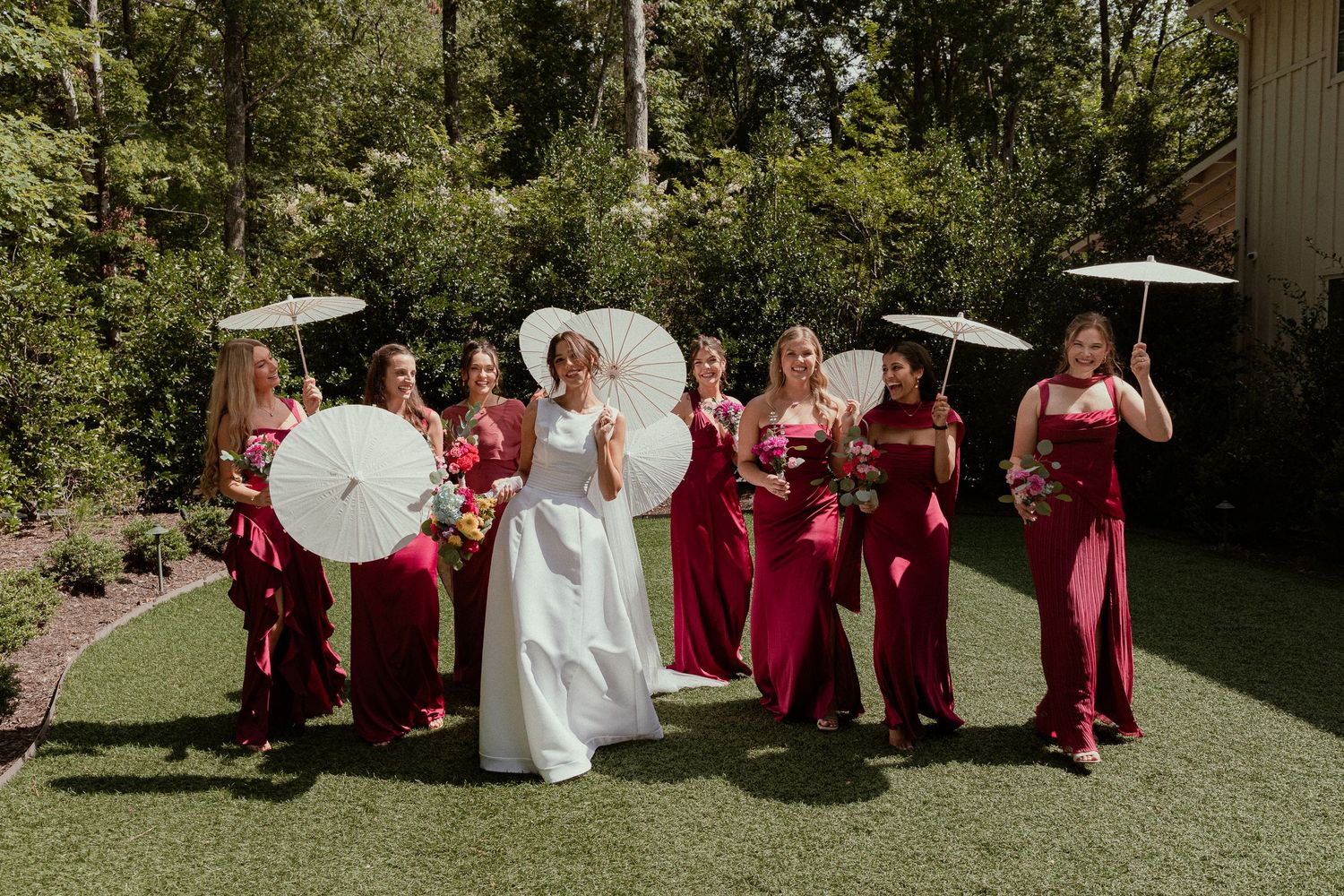 Bride in white gown holds parasol with bridesmaids in red dresses carrying white parasols outdoors.