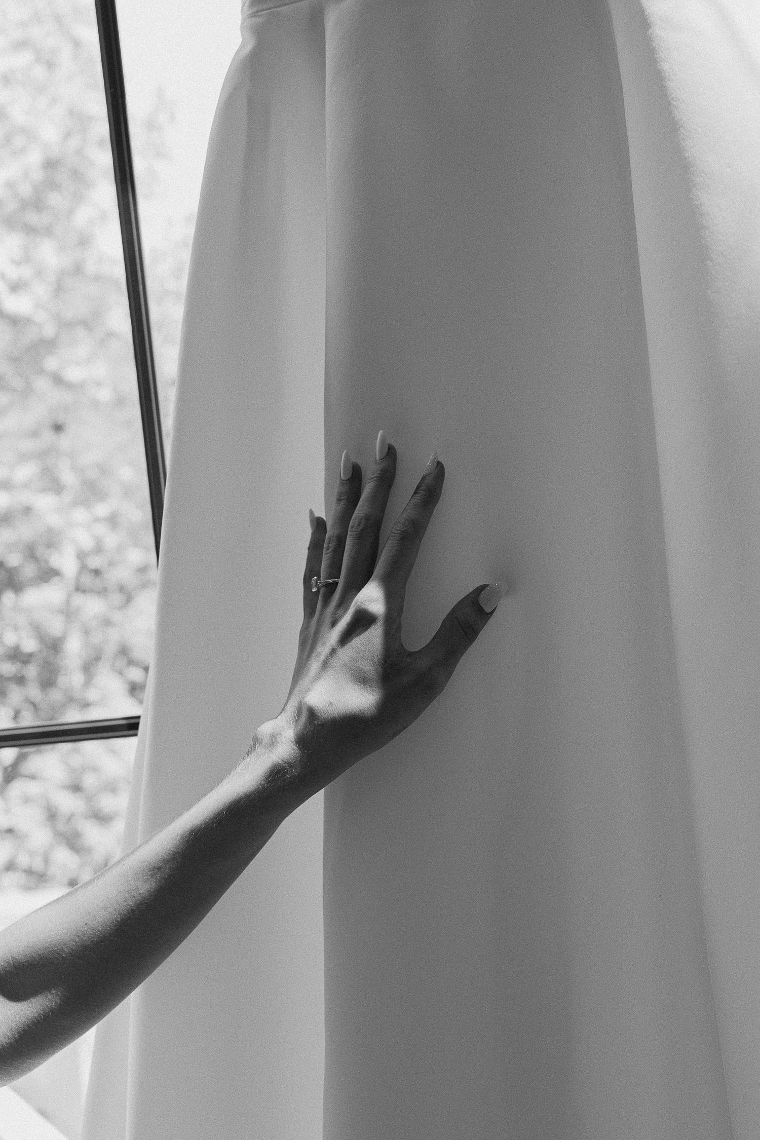 Black and white photo of a bride's hand with engagement ring touching her wedding dress near a window.