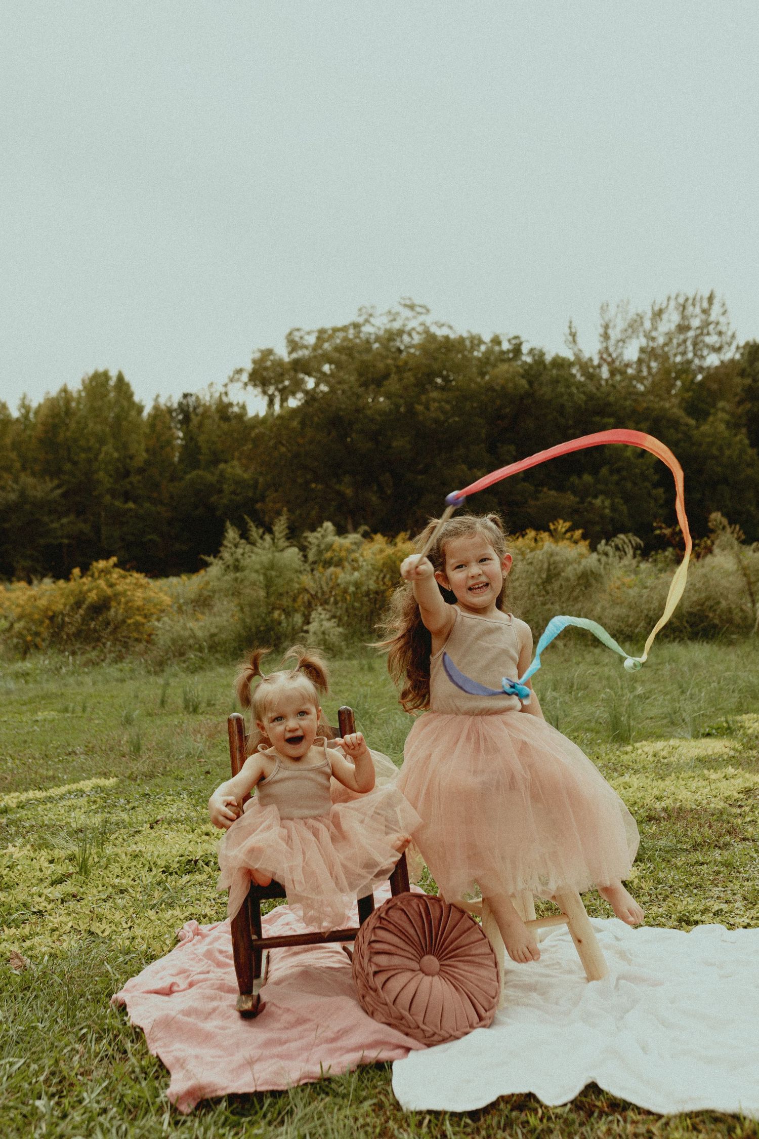 Two children in pink tulle dresses play on a blanket in a garden setting with vintage chairs.