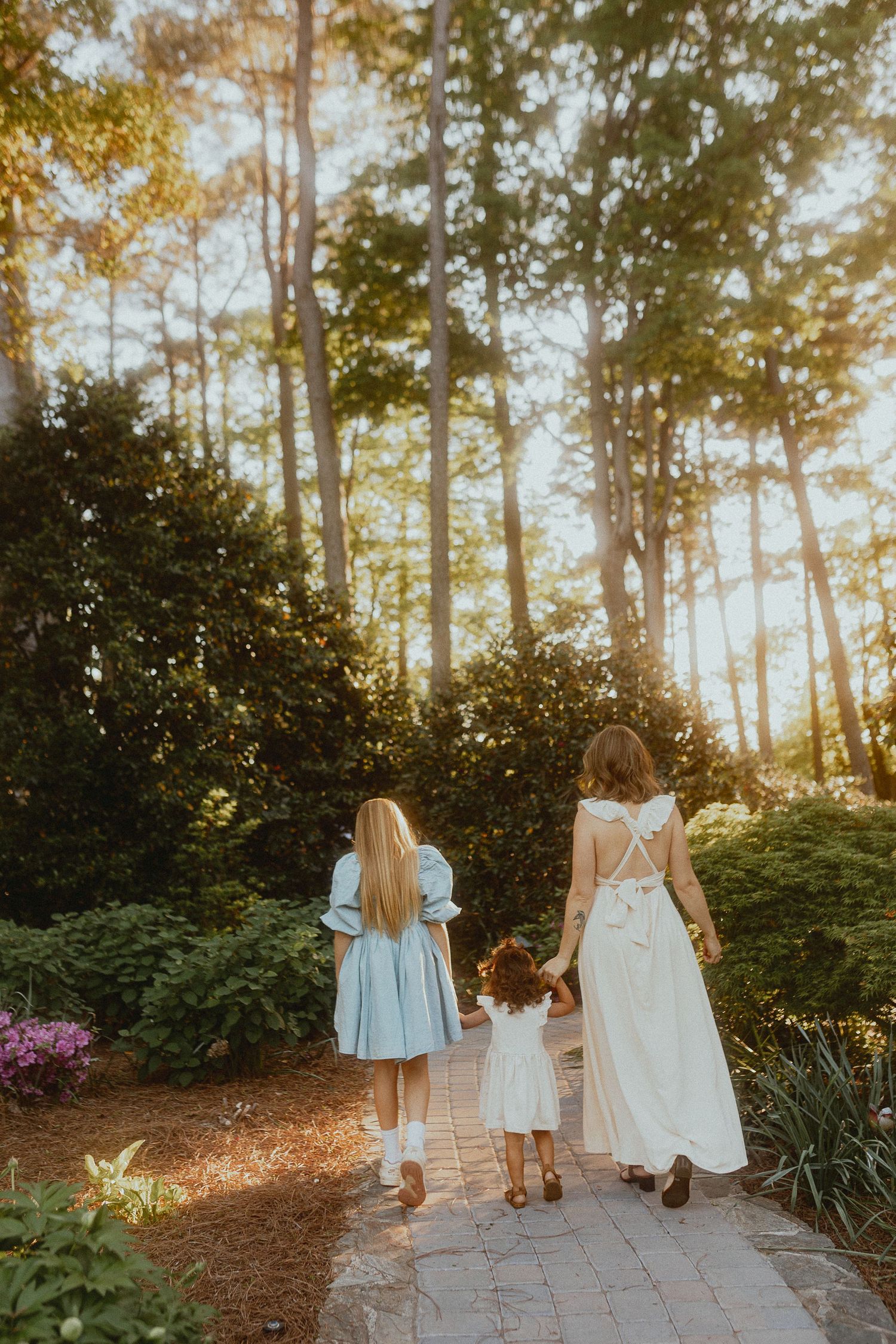 Girls in light-colored dresses walk away down a garden path surrounded by tall pine trees.