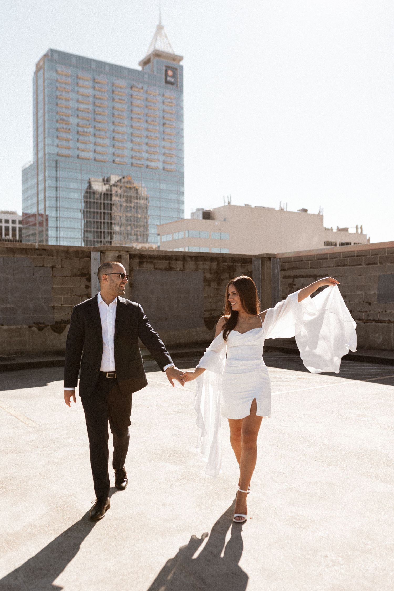 Couple in formal attire poses against urban backdrop with flowing white dress in sunlight.