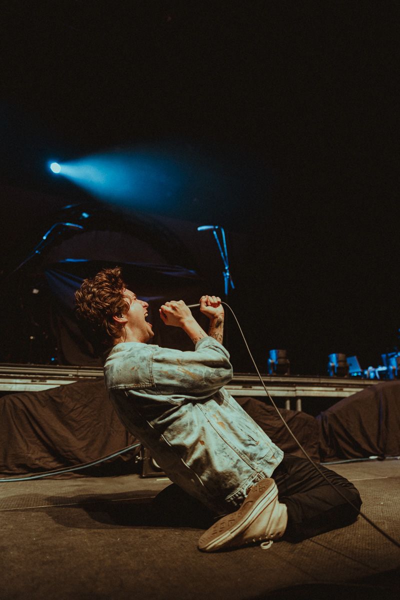 Performer kneeling on stage while singing into microphone under blue spotlight.
