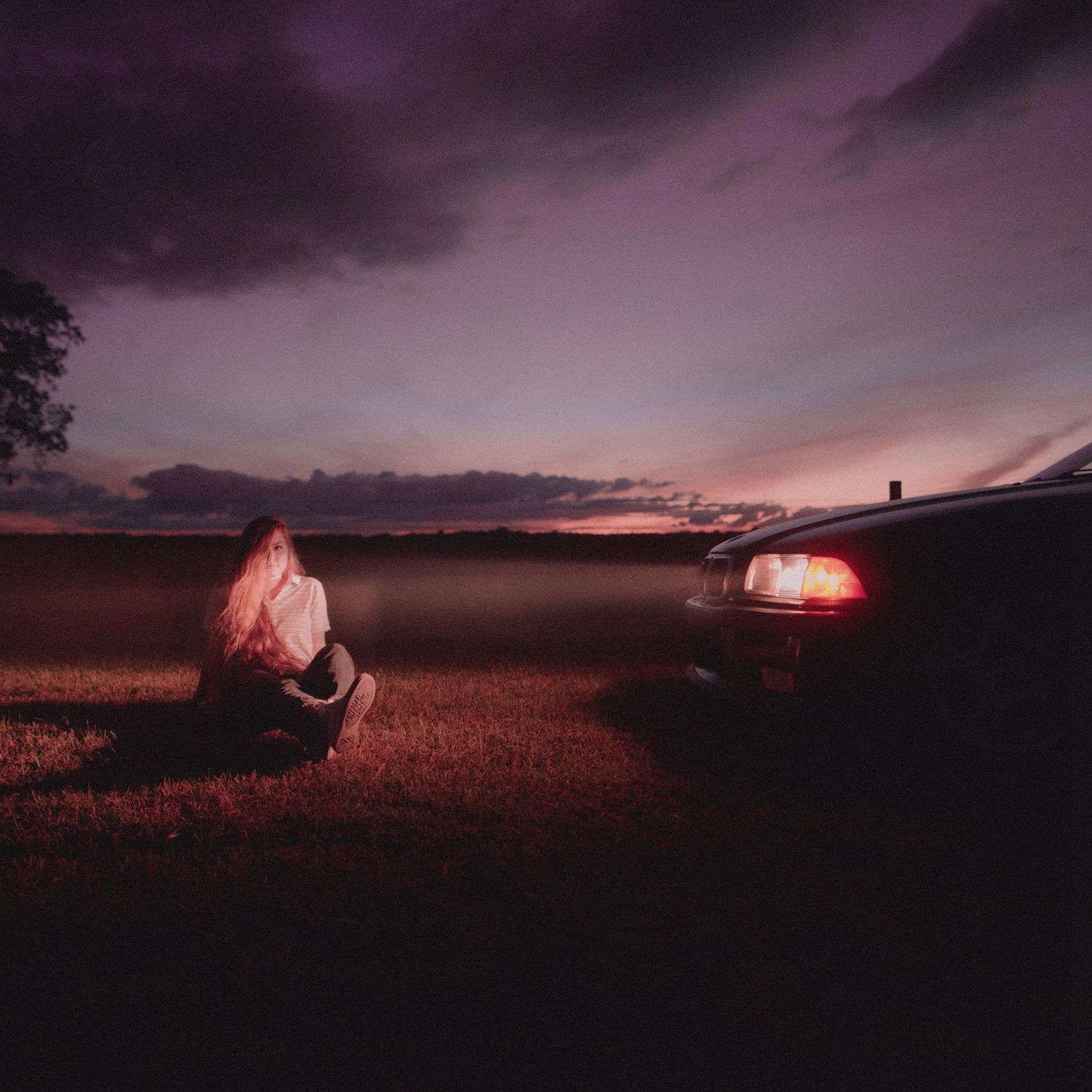 Figure sitting near illuminated tent at dusk with purple sky and dark clouds.