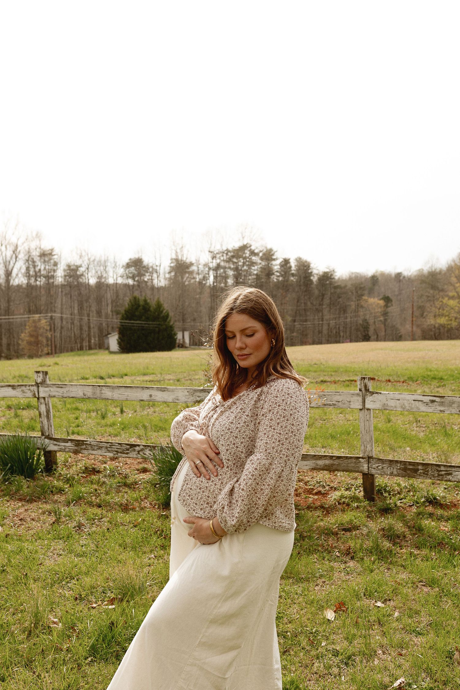 Pregnant woman in floral top and white skirt cradling her baby bump outdoors near a rustic fence.
