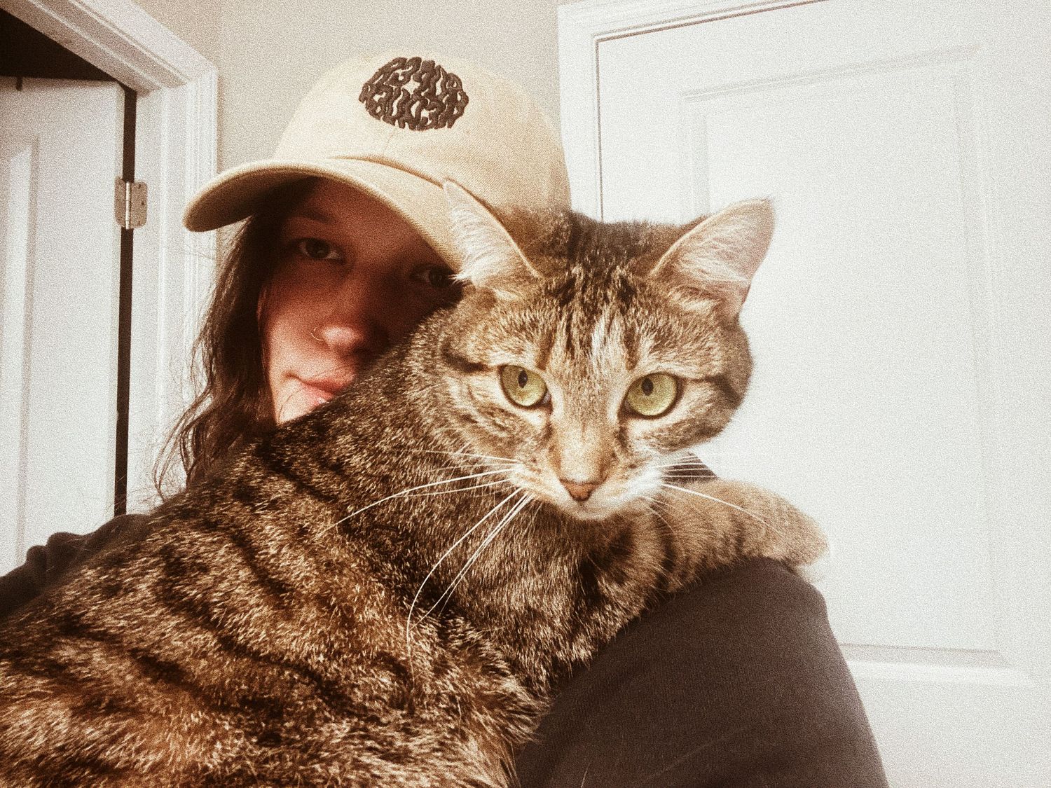 Young woman in a beige baseball cap holding a large tabby cat close for a selfie indoors.