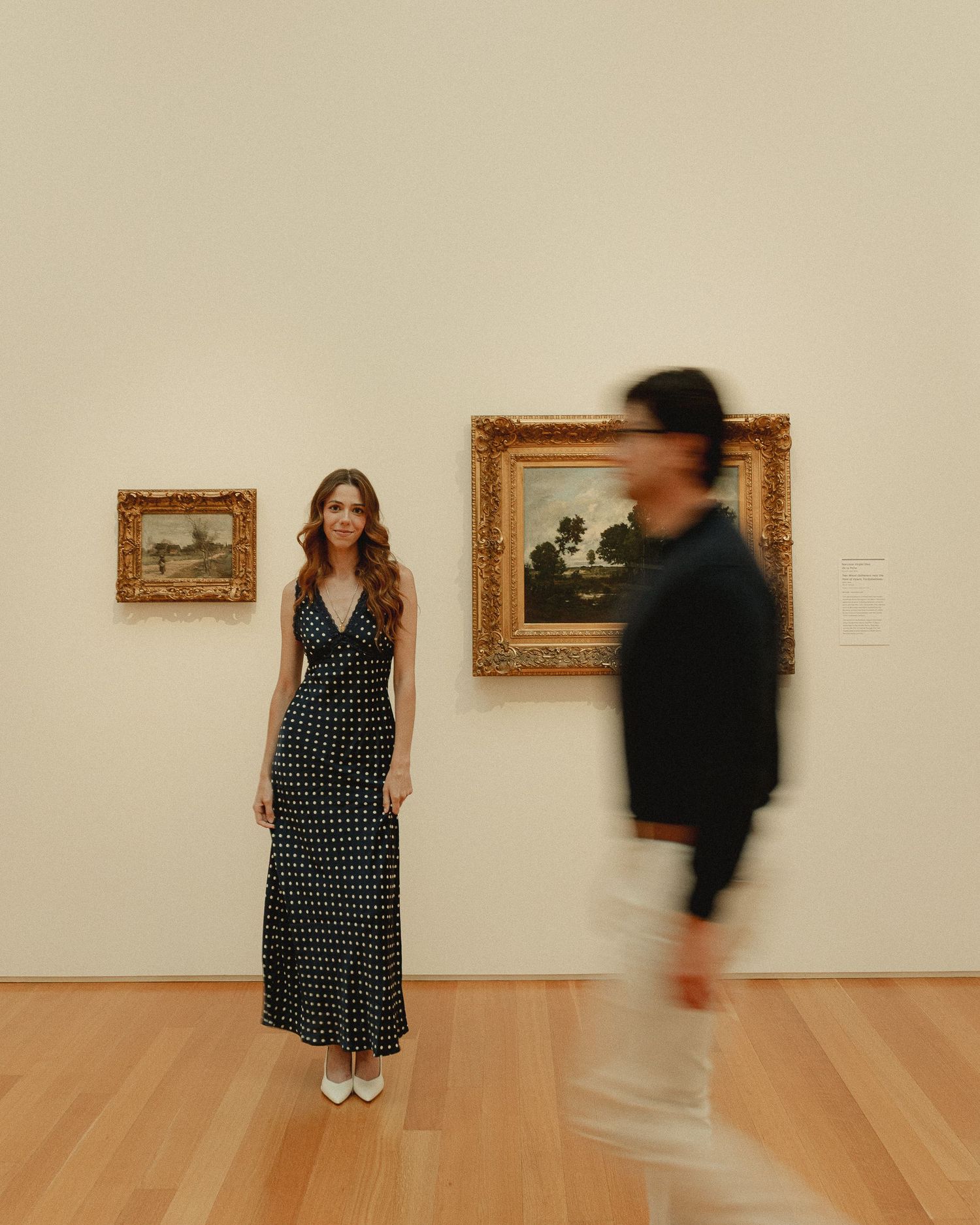 Woman in polka dot maxi dress poses in art museum gallery with gold-framed landscape paintings on wall.