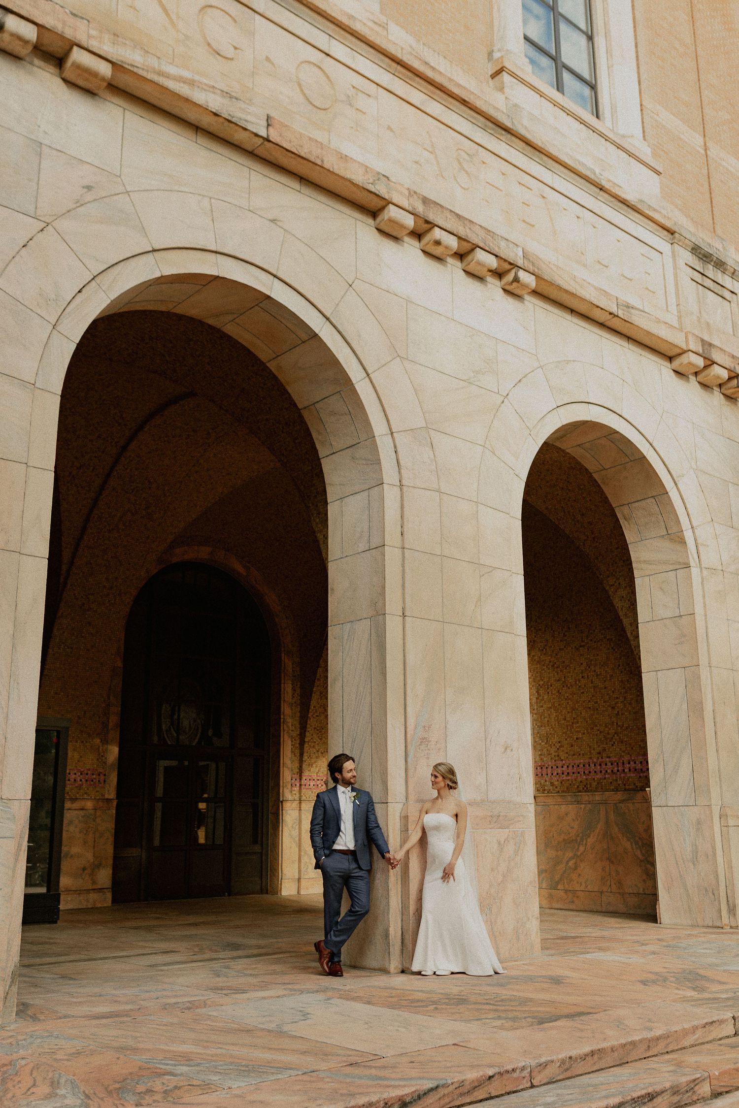 Bride and groom hold hands under elegant stone arches of a classical building in their wedding attire.