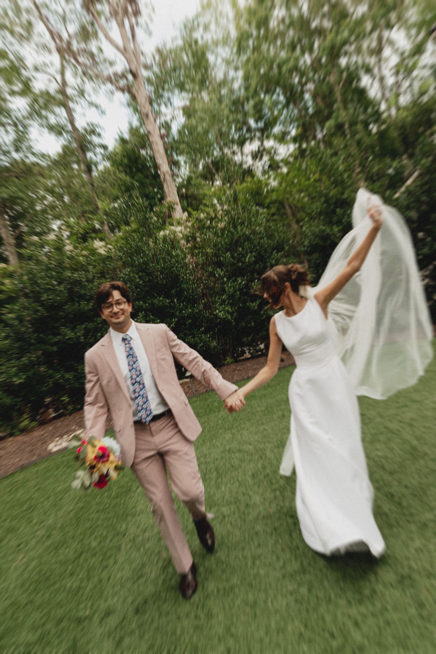 Joyful newlyweds running on grass, groom in pink suit holding bouquet, bride in white gown lifting veil outdoors.