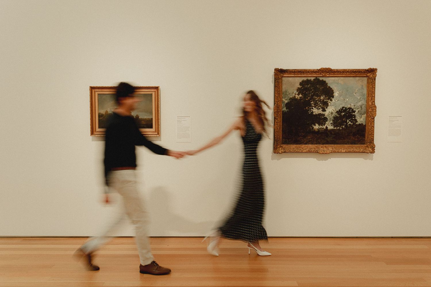 Blurred couple holding hands walking through an art museum gallery with classic paintings on white walls.