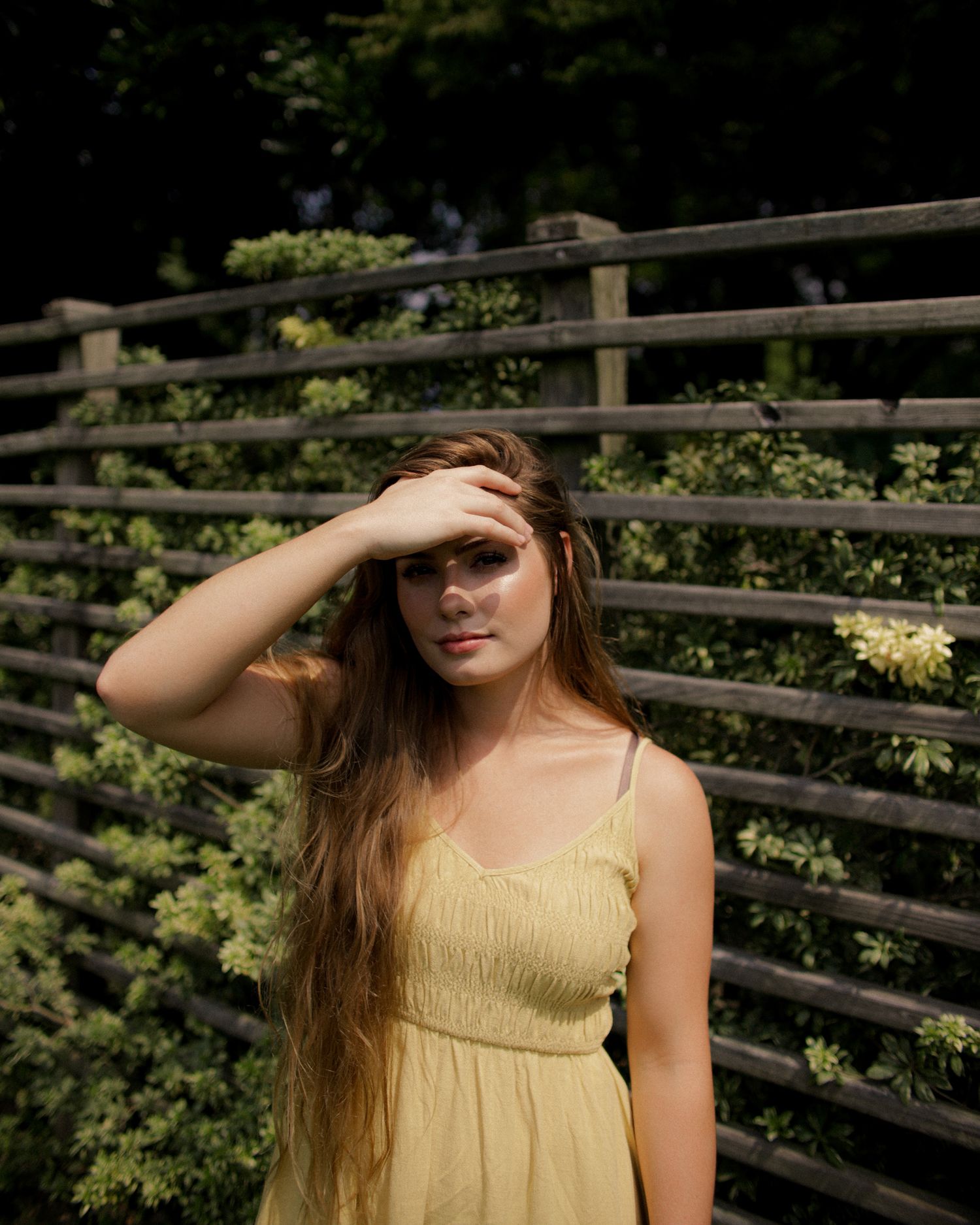 Summer portrait of someone in a yellow sundress shielding eyes from sun against a wooden fence.
