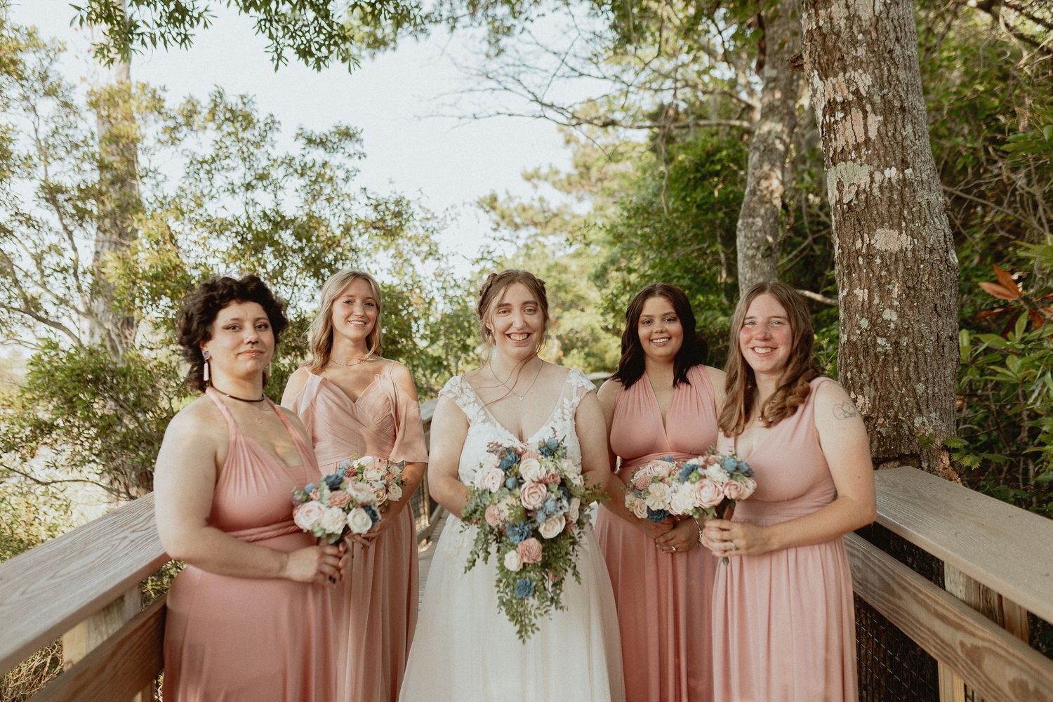 Bridesmaids in pink dresses gathered outdoors for wedding photos under trees.
