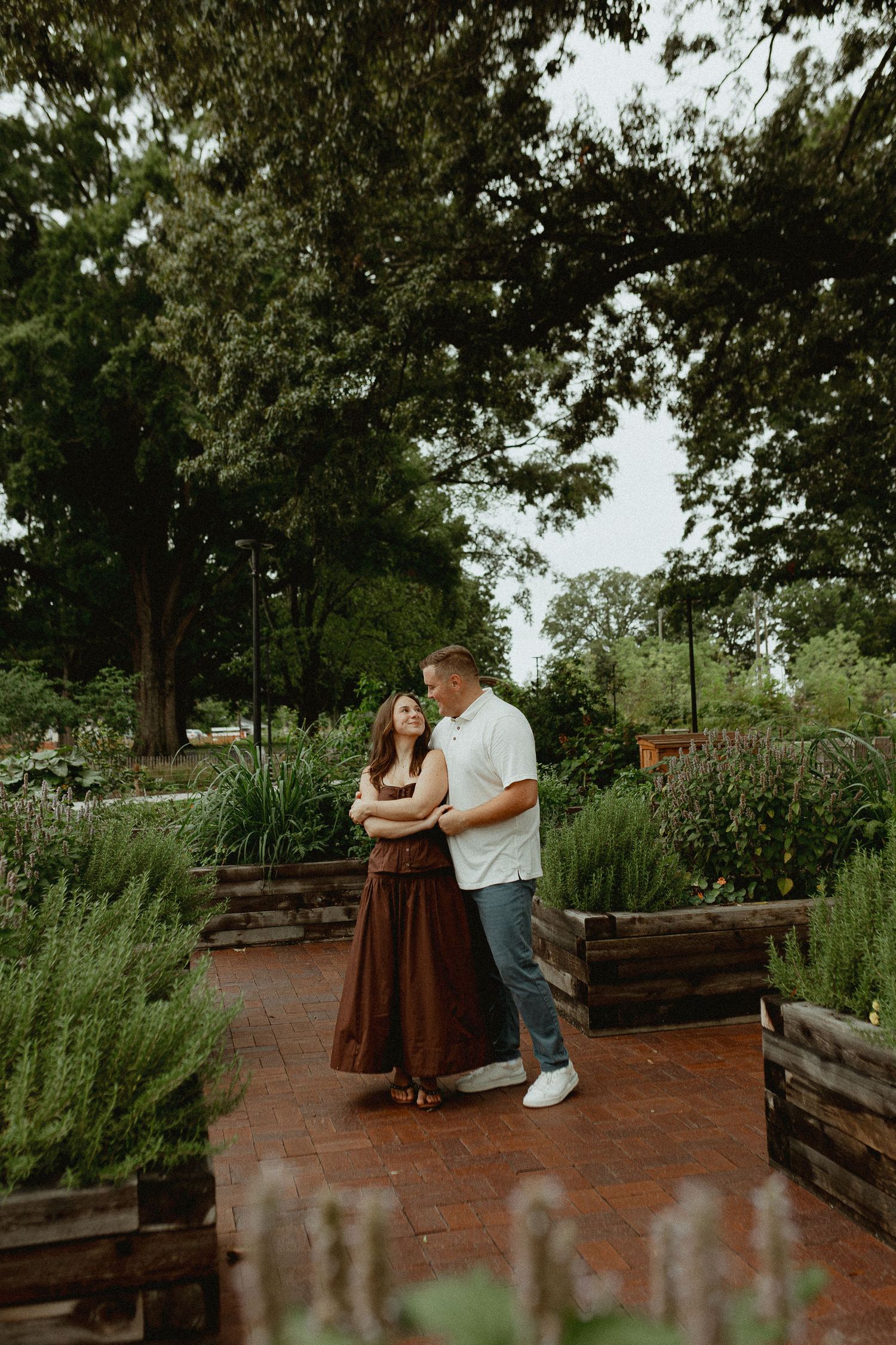 A couple shares a romantic moment on a brick path surrounded by lush garden beds and tall trees in an outdoor park.