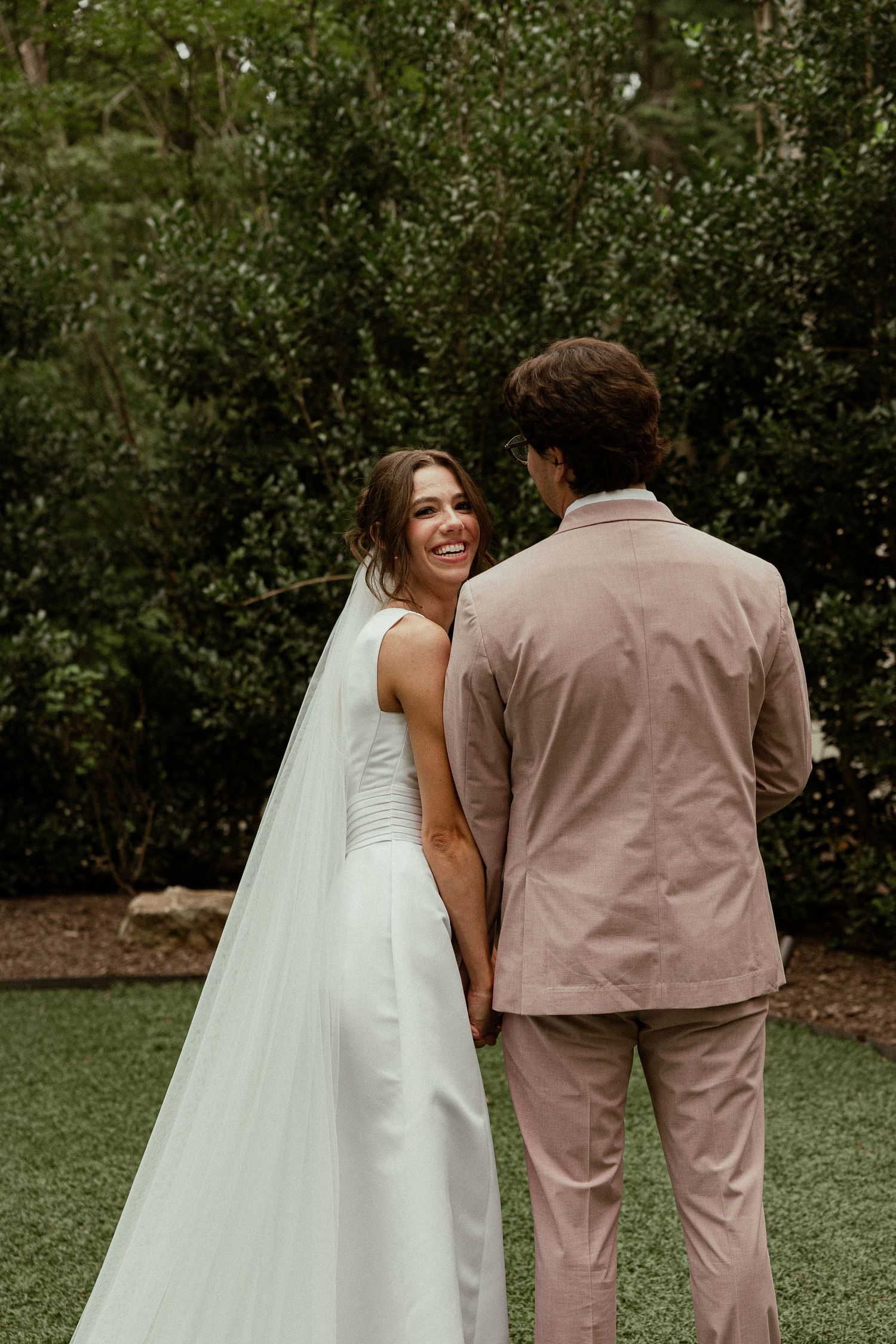 A smiling bride in a white gown and veil holds hands with a groom in a pink suit outdoors.