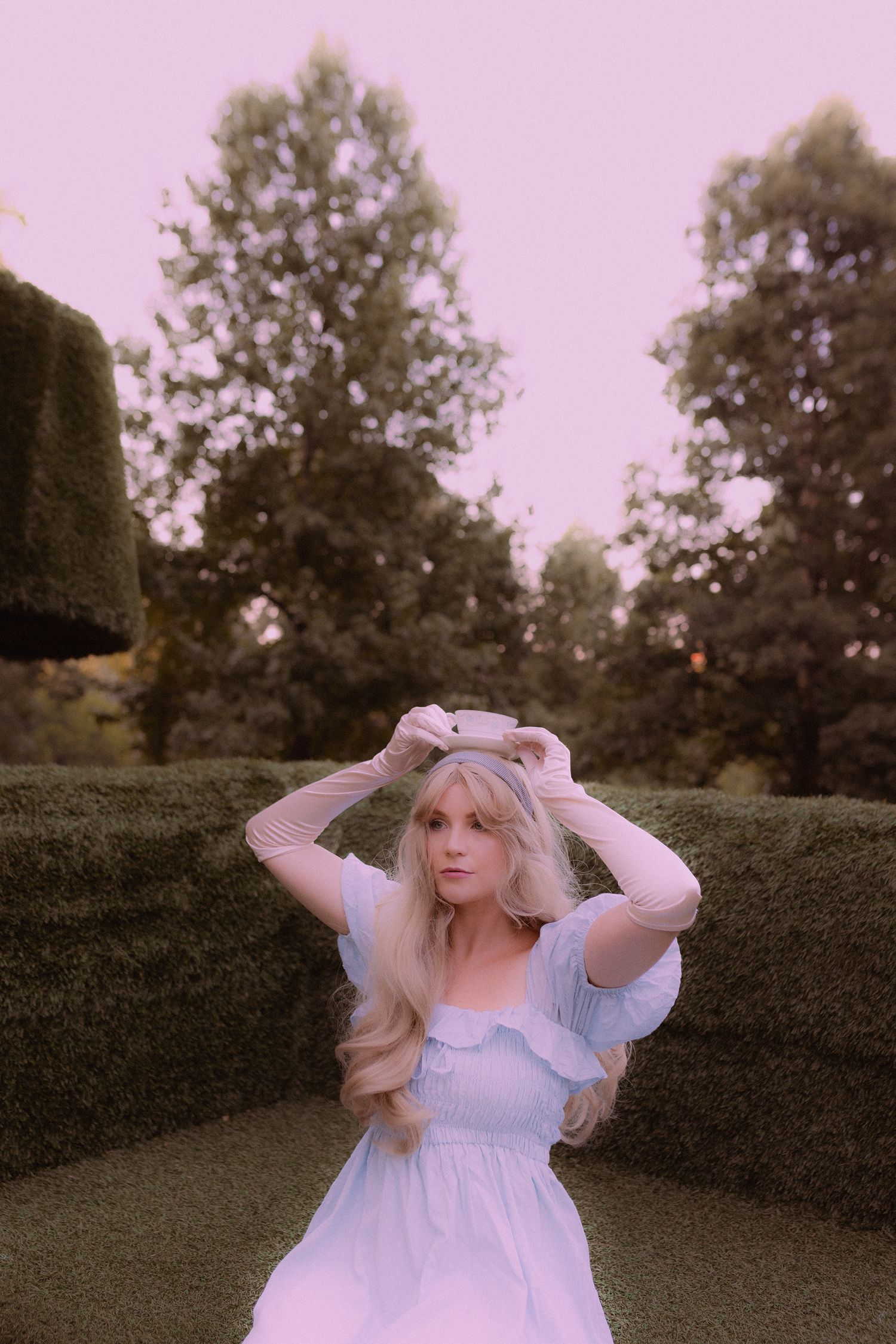 Dreamy portrait of a person in a light blue vintage dress posing in a garden with hedges at sunset.