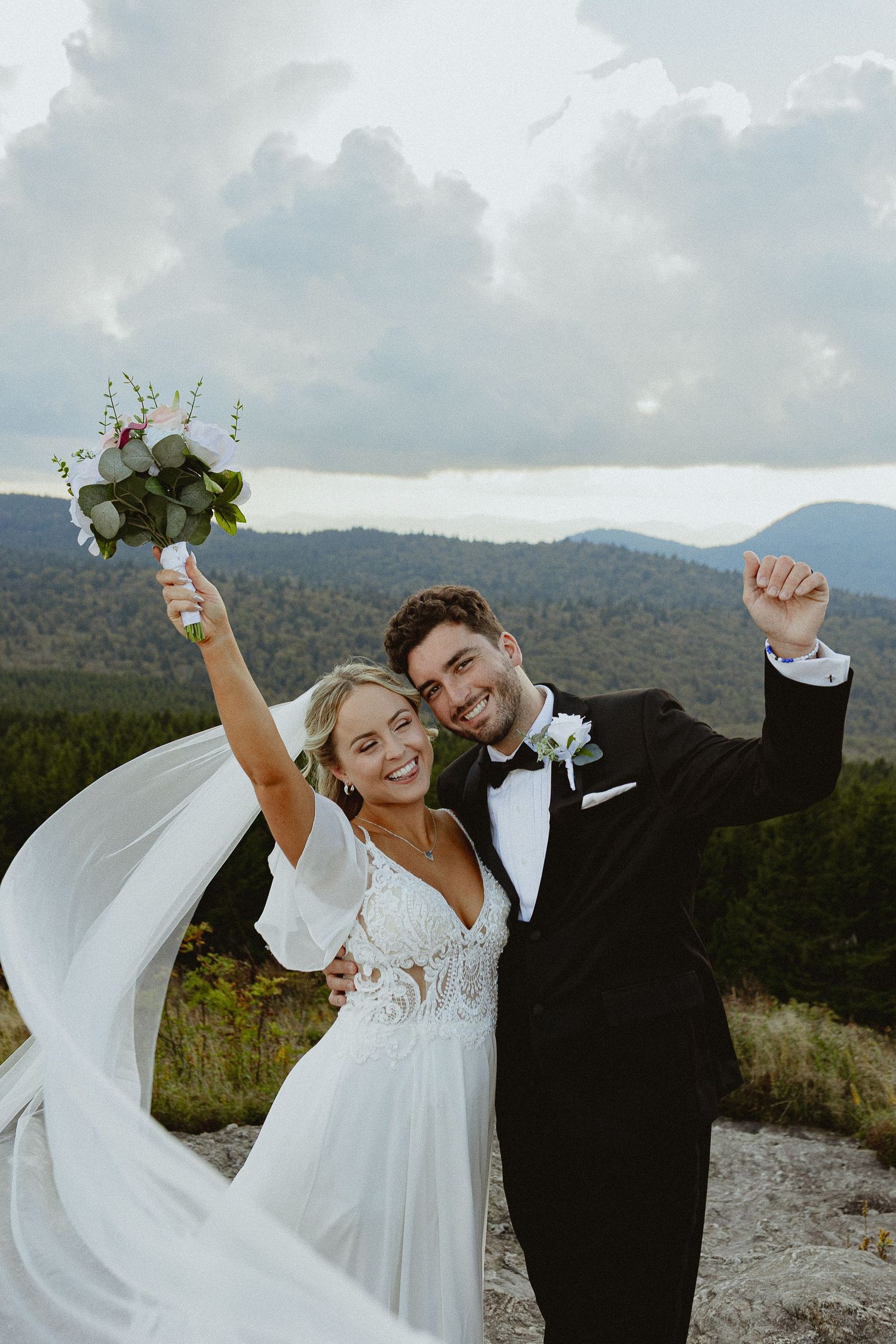 Joyful newlyweds celebrate on a mountain overlook, bride in white gown holding bouquet, groom in black tuxedo.